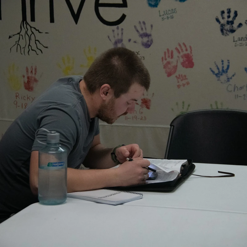 A young man with a beard sits at a white table, studying from a book and a phone. The wall behind him is covered in colorful children's handprints.