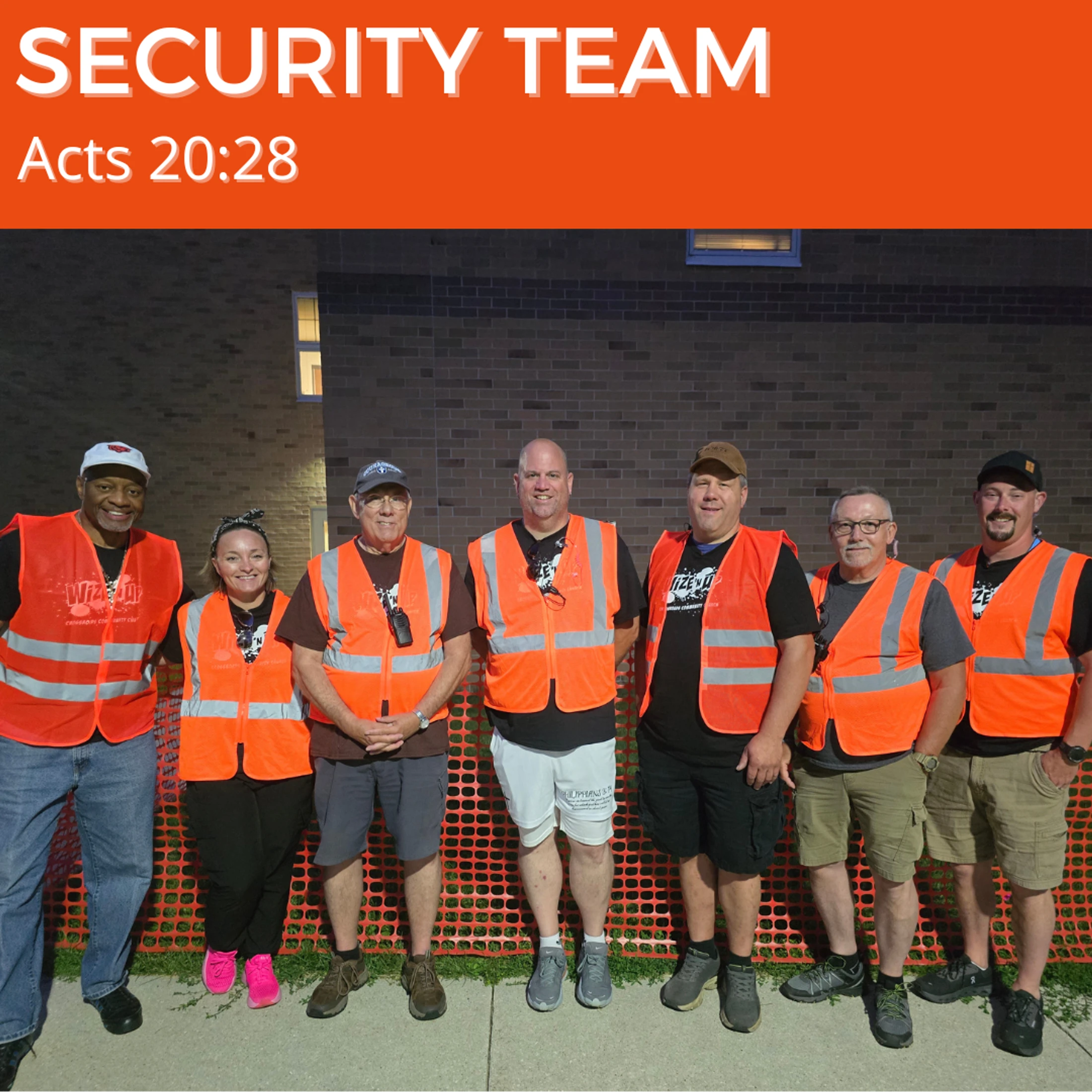Seven security team members, six men and one woman, stand in a line smiling for a photo at night. They are all wearing orange safety vests.