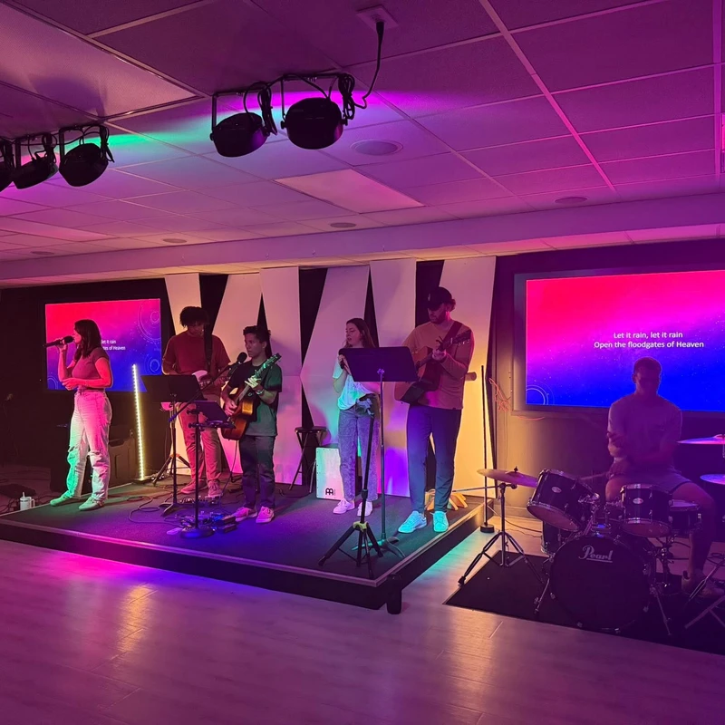 A band with singers, guitarists, a keyboardist, and a drummer performs on a stage bathed in pink and purple light, with lyrics displayed on screens.