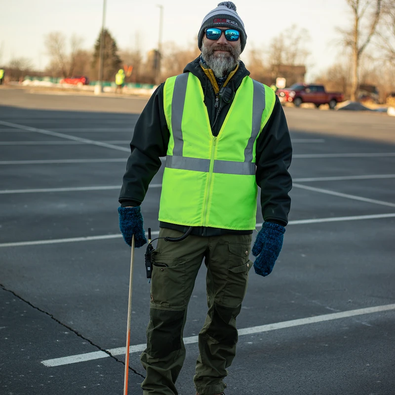 A bearded man wearing a winter hat, sunglasses, and a bright yellow safety vest stands in an empty parking lot while holding a small directional pole.