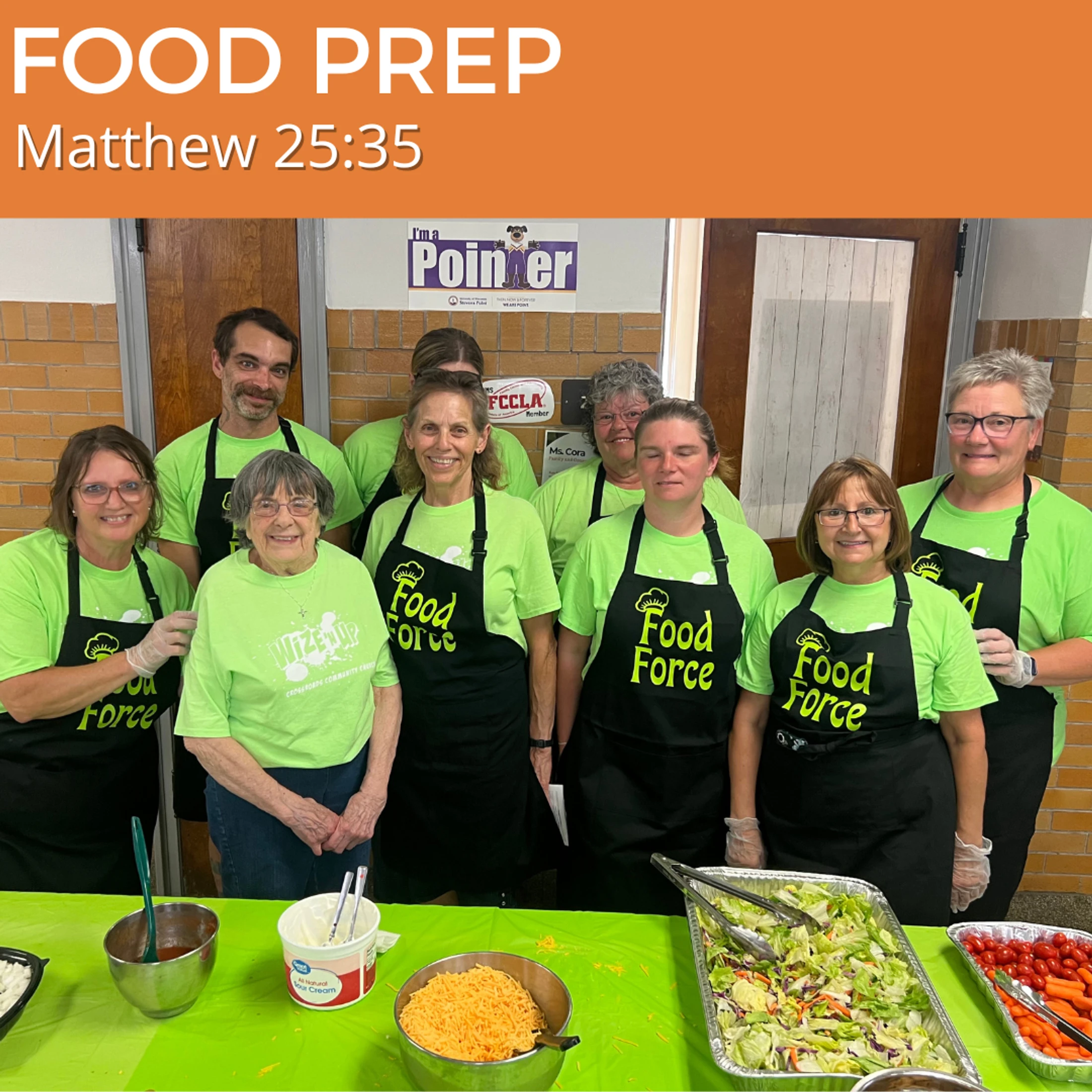 A group of smiling volunteers in matching 'Food Force' aprons and shirts stand behind a table of food ingredients ready to serve a meal.