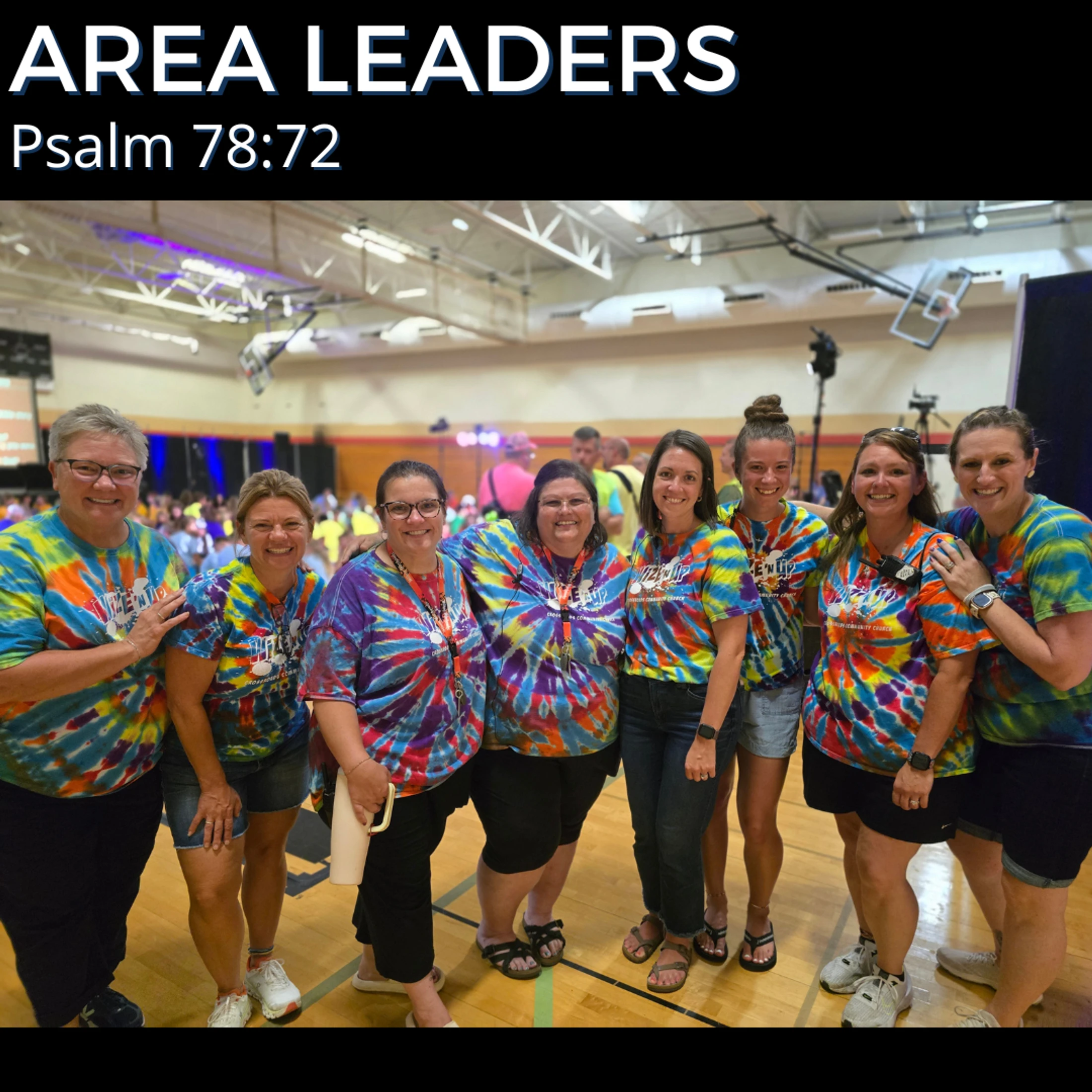 A group of eight smiling women, the Area Leaders, wearing colorful matching tie-dye shirts pose for a photo in a gymnasium during an event.