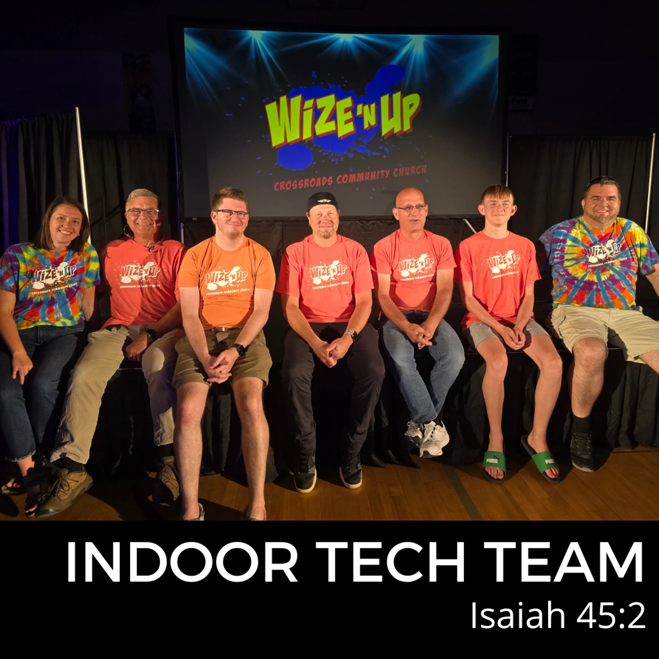 A group of seven people, the Indoor Tech Team, sit on a stage smiling for a photo. They wear t-shirts for a 'Wize 'n Up' church event.