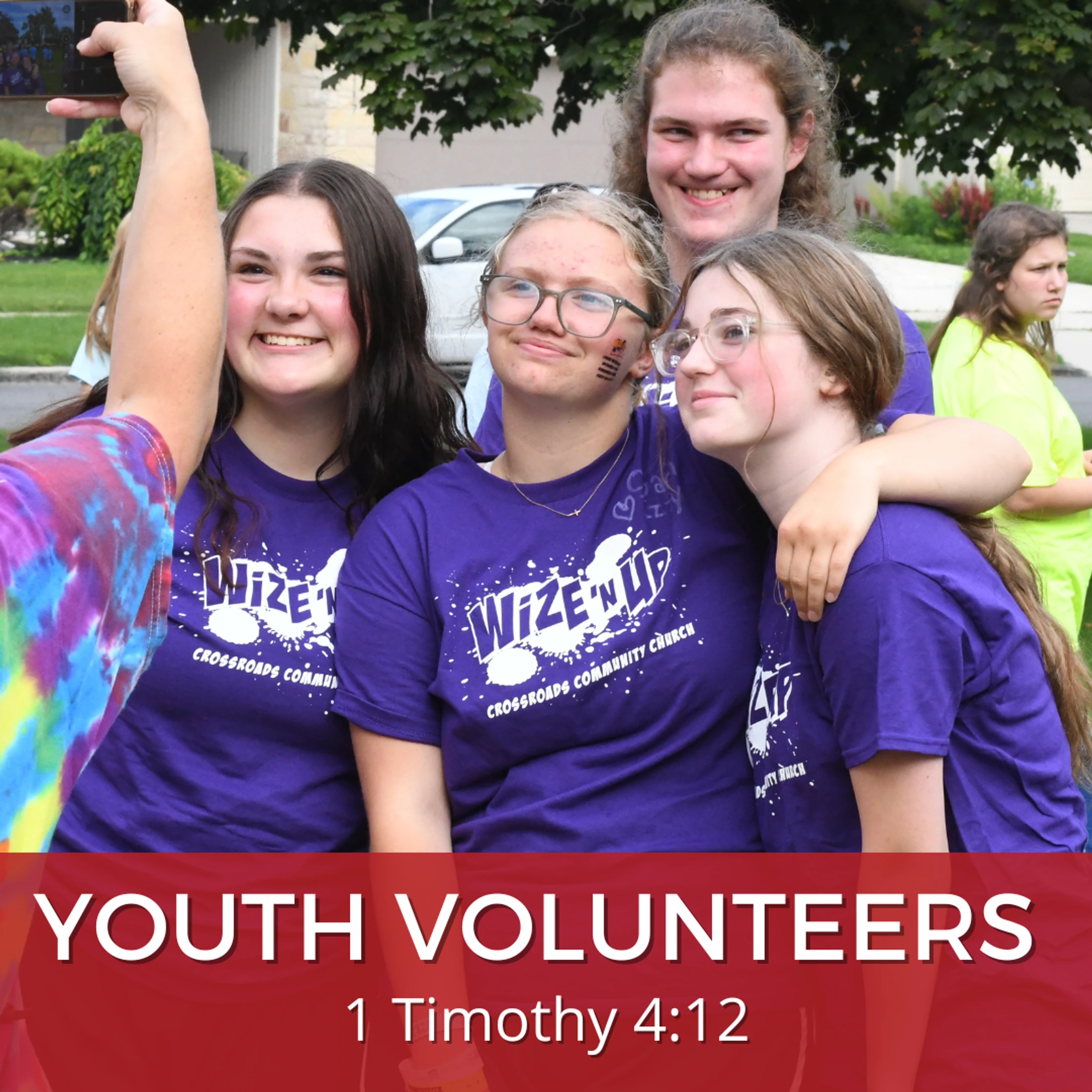 A group of four smiling young volunteers in matching purple church t-shirts pose for a selfie being taken by a person in a tie-dye shirt.