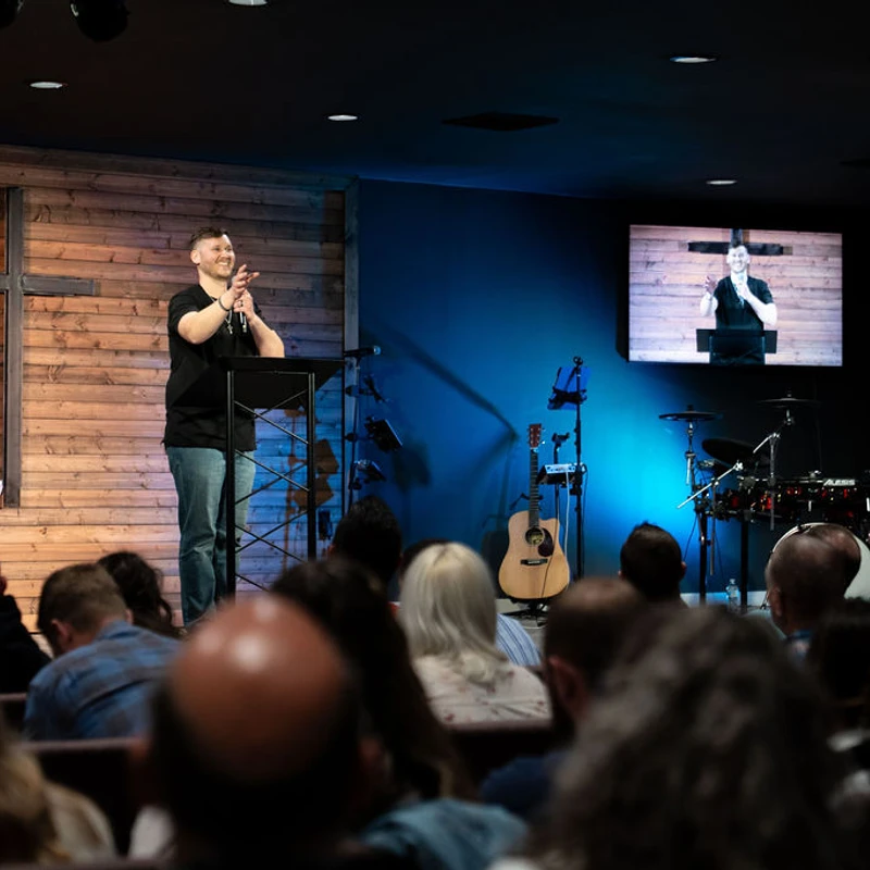 A man stands at a lectern on a stage with a wooden wall and a cross, speaking to an audience. A screen shows him, and musical instruments are nearby.