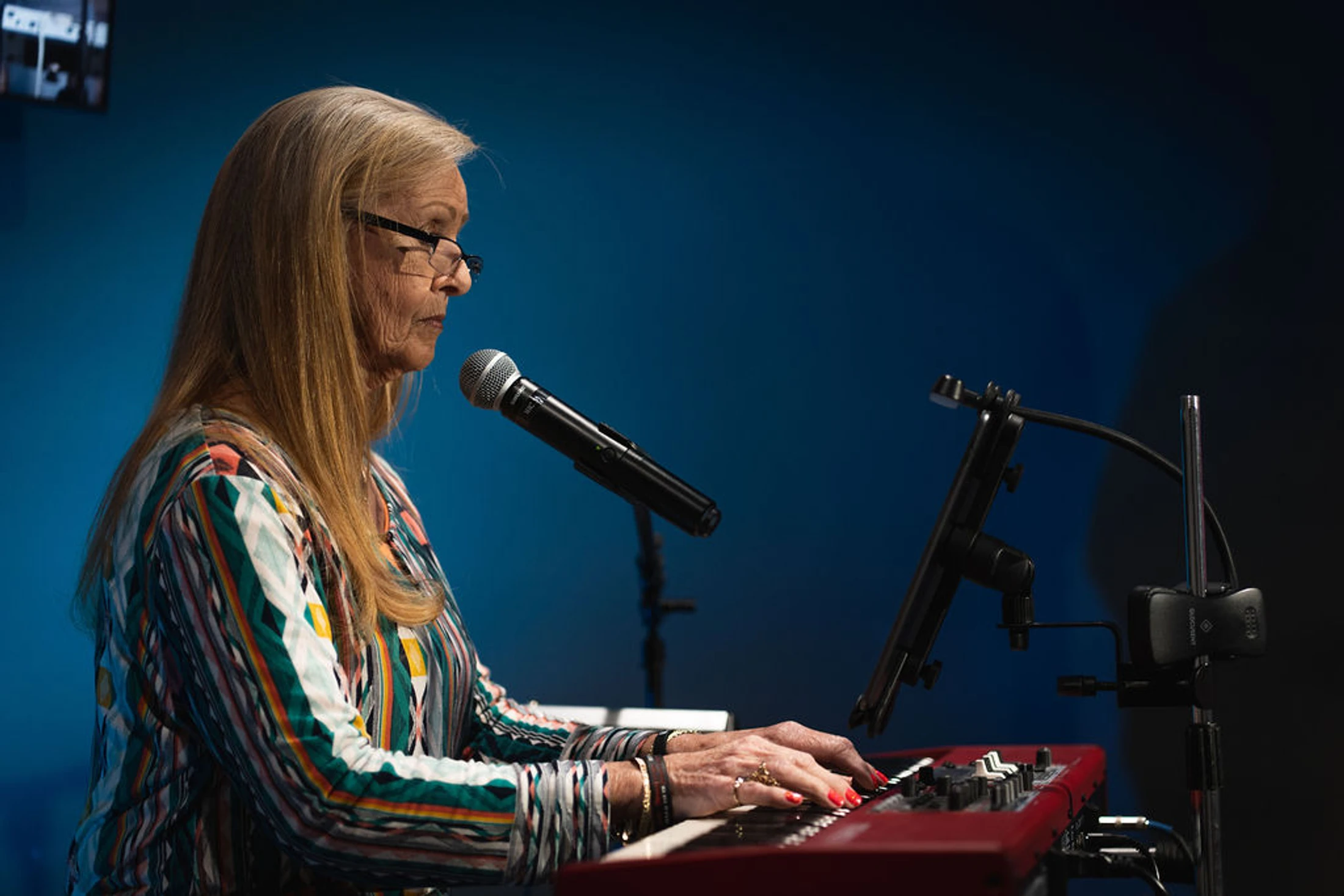 A side-profile view of a woman with long hair and glasses playing a red keyboard. A microphone is positioned in front of her against a blue background.