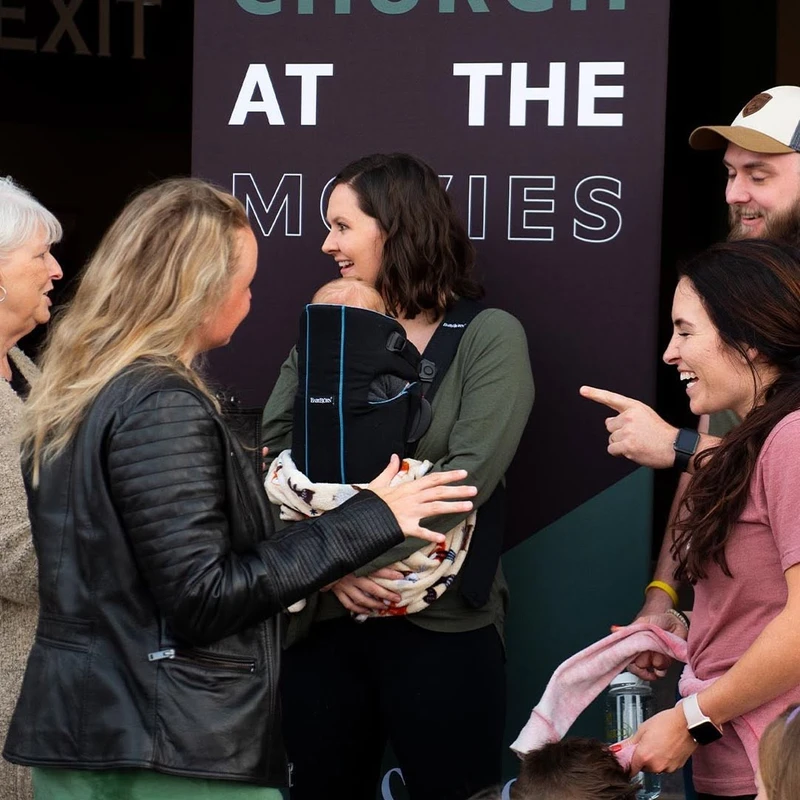 A group of people smiles and talks together, with a woman in the center holding a baby in a carrier, in front of a "Church at the Movies" banner.