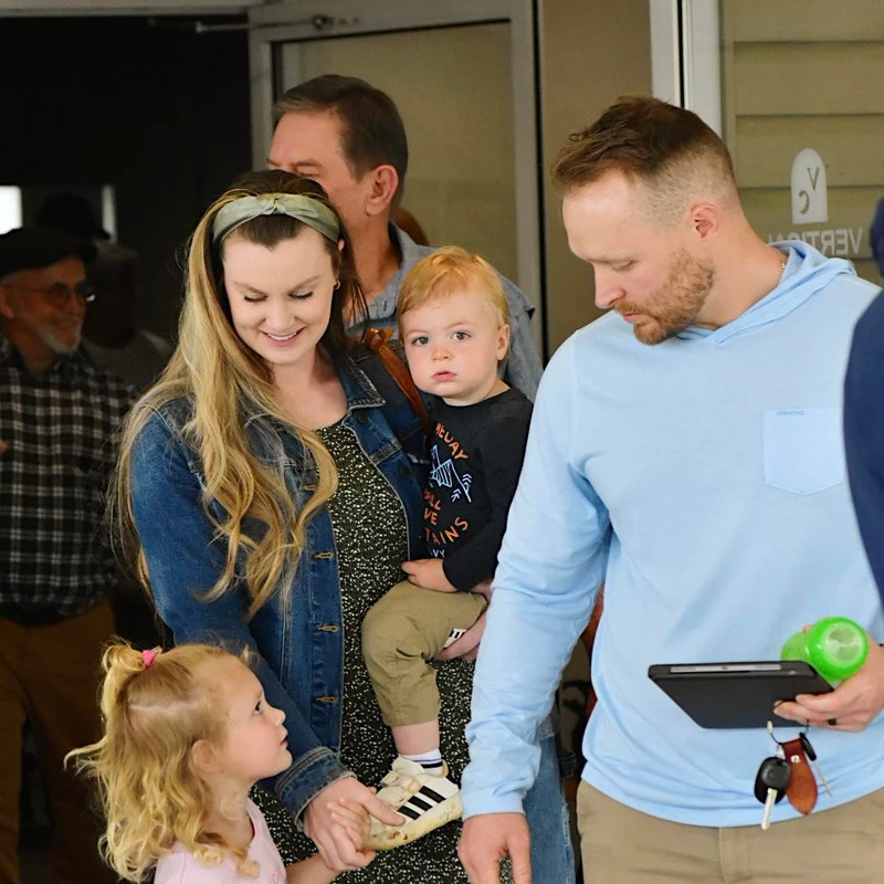A family stands together indoors. A woman in a jean jacket holds a toddler, while a little girl looks up at her. A man in a blue hoodie stands beside them.