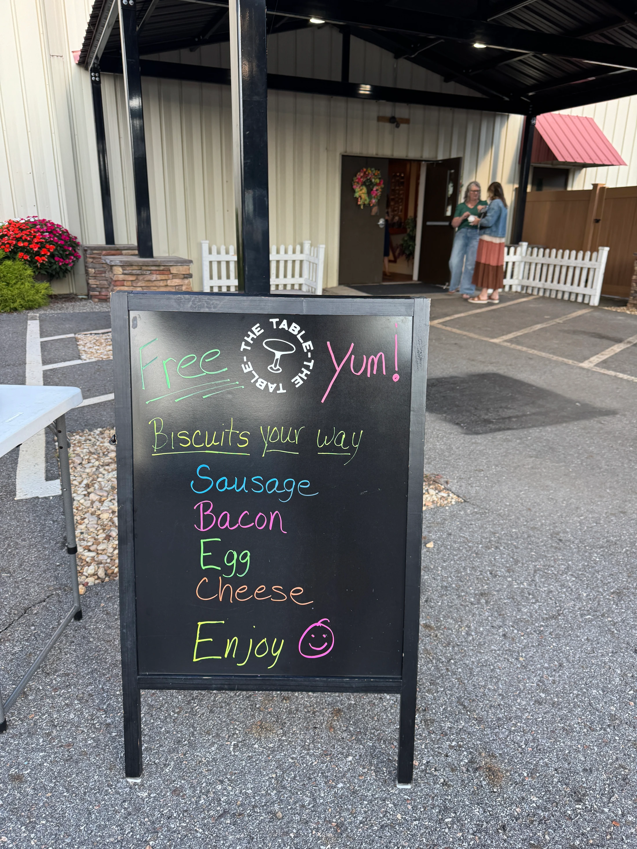 A colorful chalkboard A-frame sign on pavement advertises free biscuits with toppings like sausage, bacon, egg, and cheese outside a cafe called The Table.