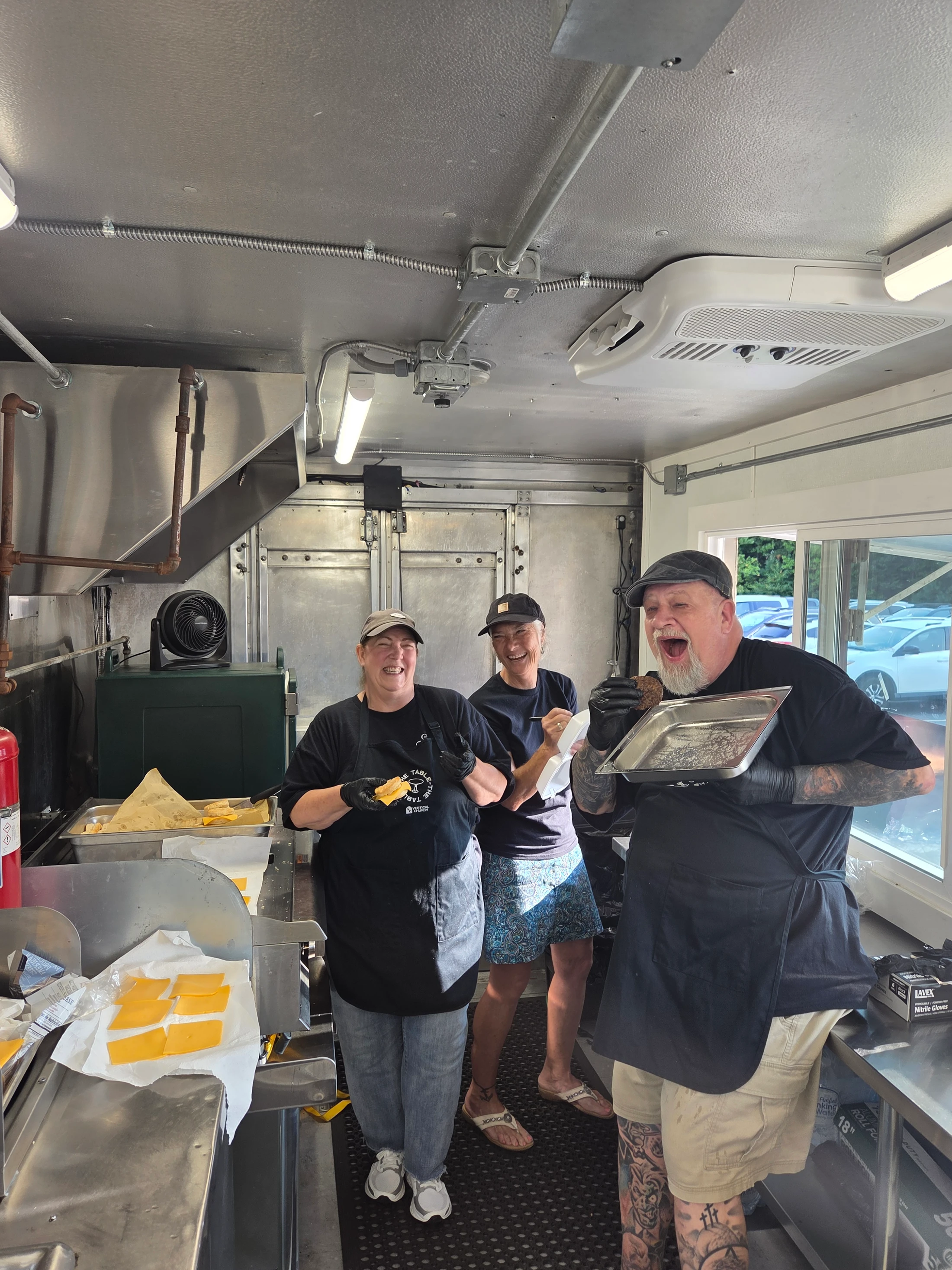 Three smiling staff members pose inside a food truck. A woman holds a sandwich, while a tattooed man on the right playfully shouts with his mouth open.