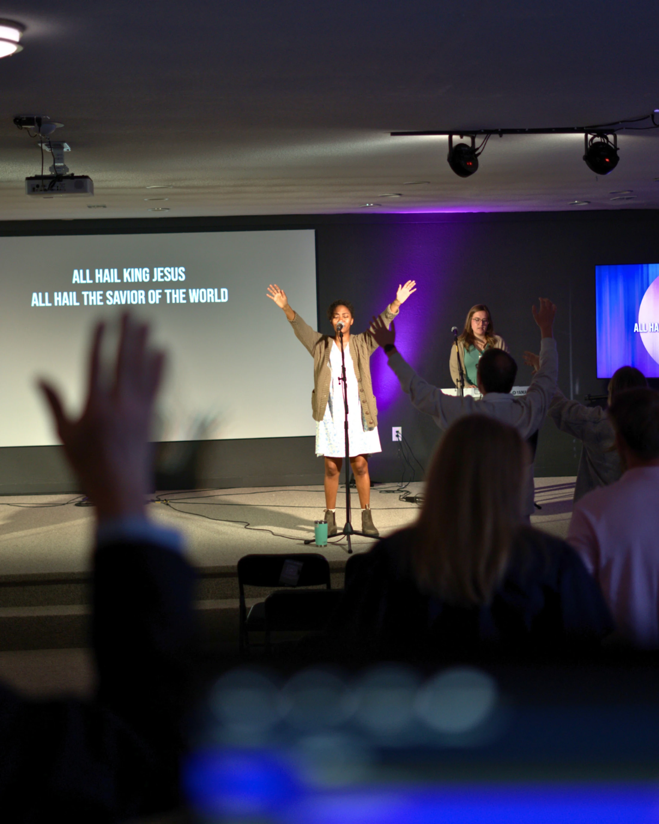 From the audience's view, a woman leads a worship song on stage with her arms raised. A screen behind her displays the lyrics 'ALL HAIL KING JESUS'.