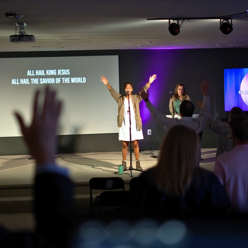 From the audience's view, a woman leads a worship song on stage with her arms raised. A screen behind her displays the lyrics 'ALL HAIL KING JESUS'.