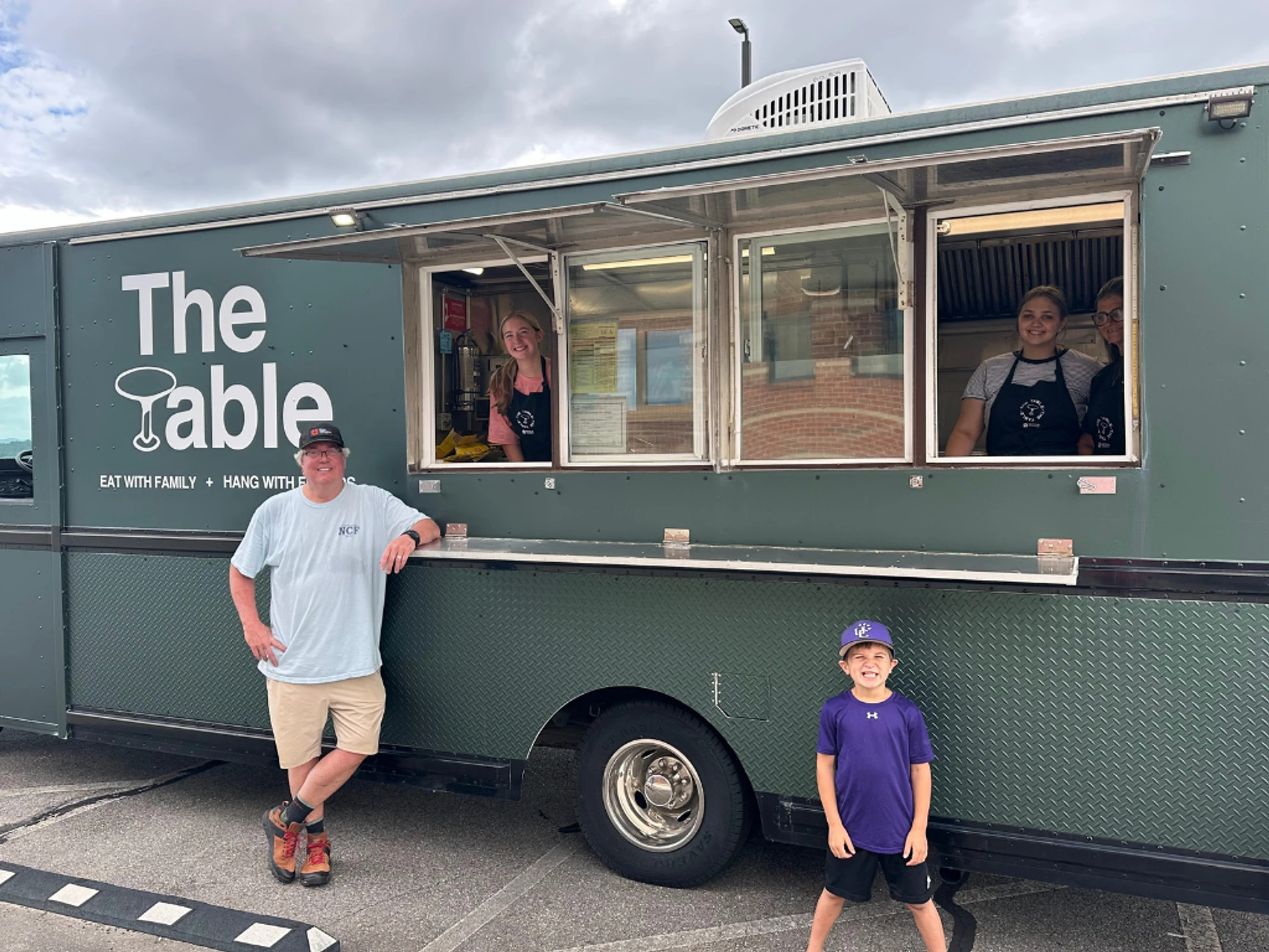 A man and a young boy stand outside a green food truck named 'The Table' as three female employees smile from the service window.