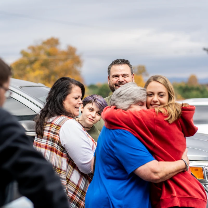 A group of people smile while watching two women share a tight, warm hug outdoors on a cloudy day, with a car and autumn trees in the background.