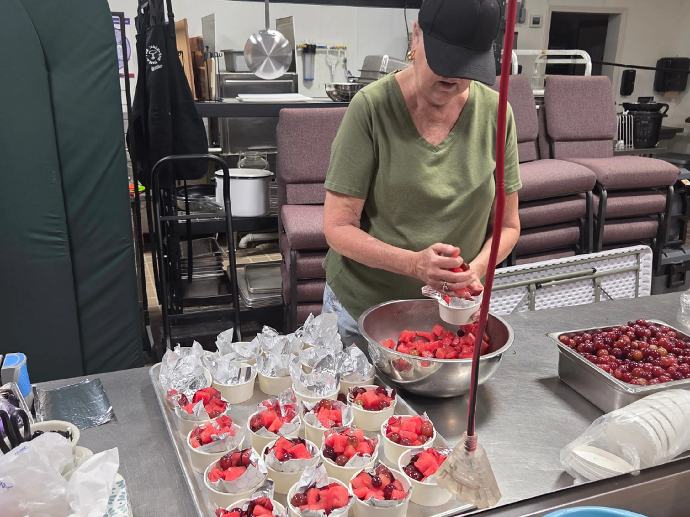 A woman in a green shirt and black cap prepares individual fruit cups with watermelon and grapes at a stainless steel counter in a commercial kitchen.
