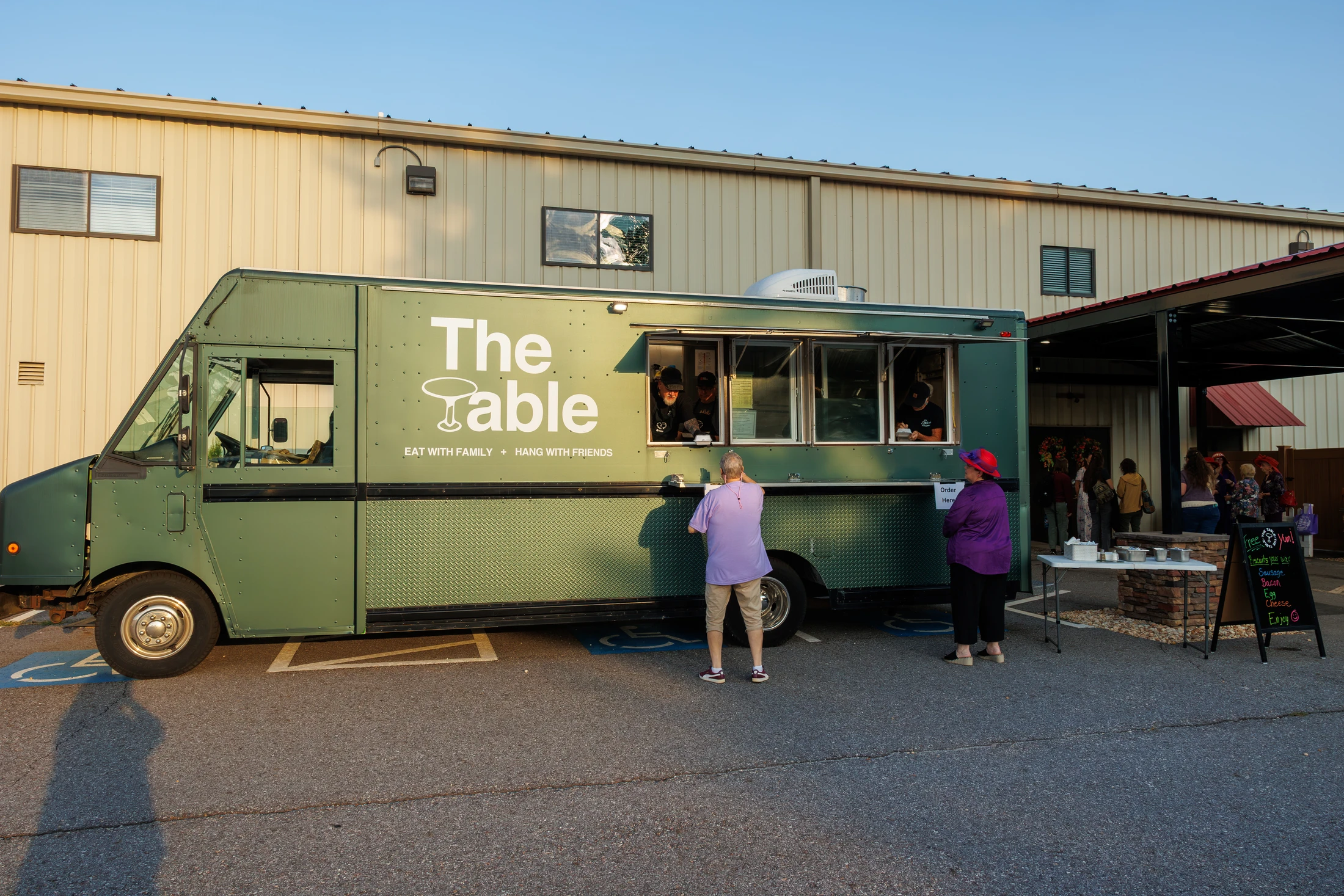 A green food truck named 'The Table' is parked in a lot, with several customers waiting in line to order from the service window during an evening event.