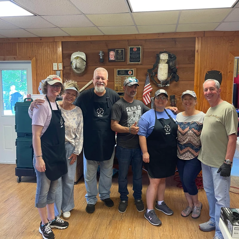 A group of seven smiling volunteers, men and women in aprons and casual wear, pose for a photo inside a room with wood-paneled walls and firefighter decor.