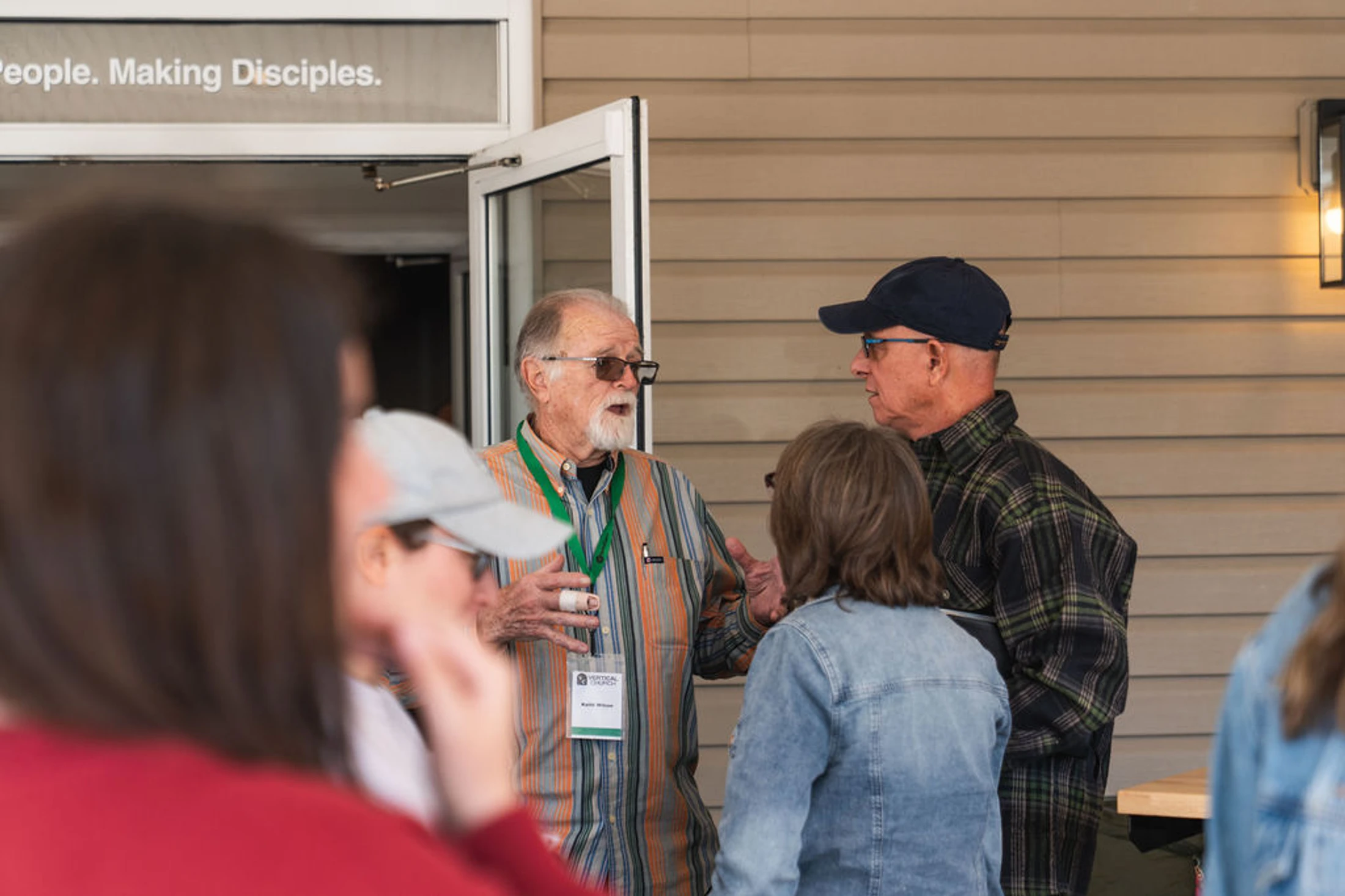 An older man with a goatee gestures while speaking to a small group outside a building with a sign above the door reading 'People. Making Disciples.'