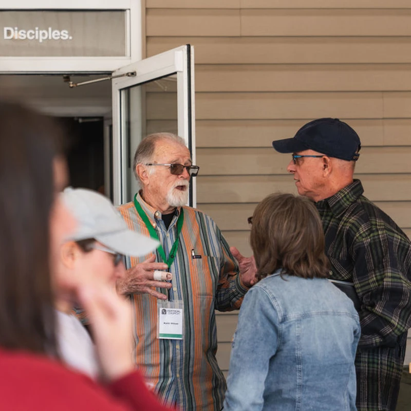 An older man with a goatee gestures while speaking to a small group outside a building with a sign above the door reading 'People. Making Disciples.'