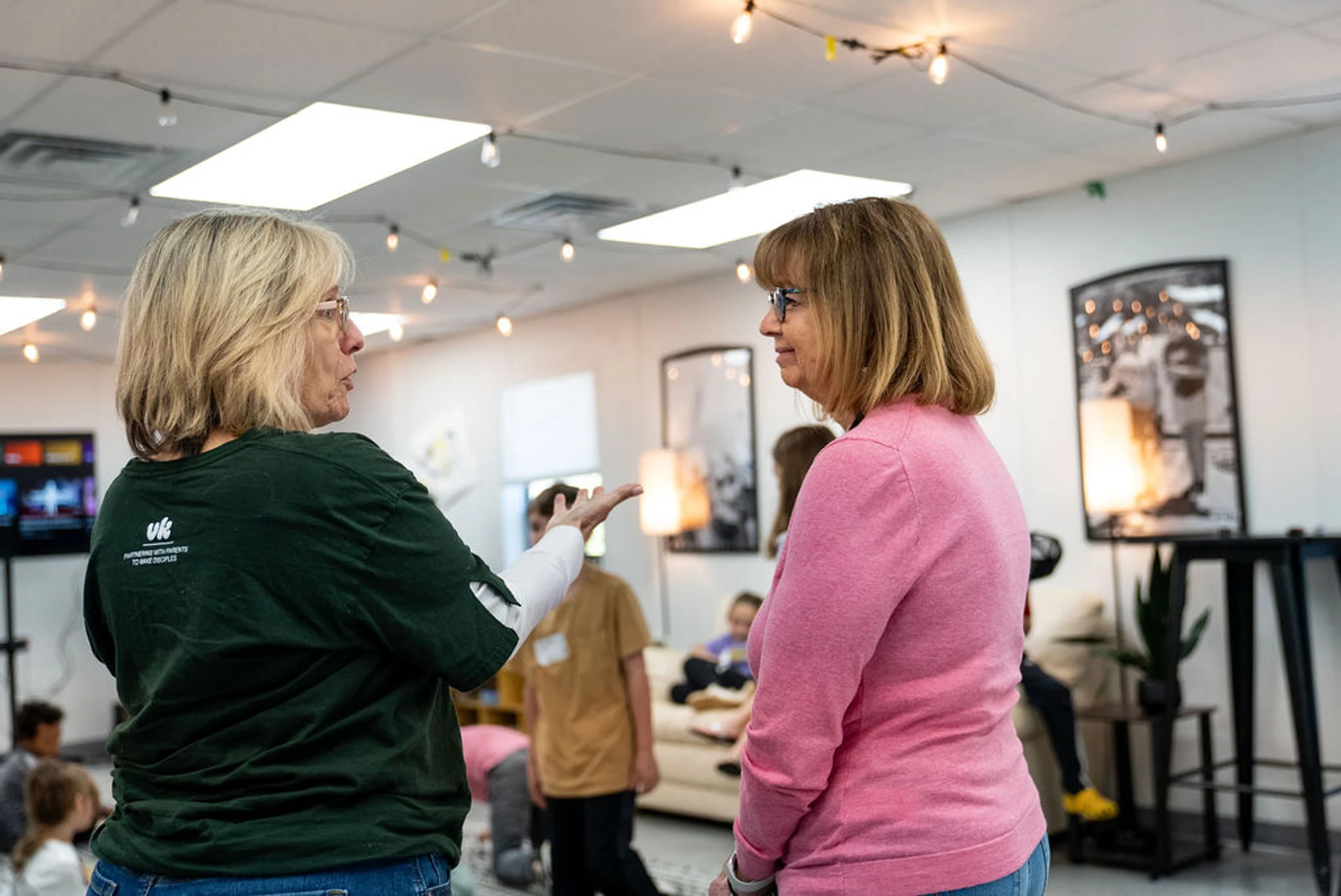 Two women stand talking in a well-lit room. One woman in a green t-shirt gestures as she speaks to another woman in a pink sweater who is listening intently.