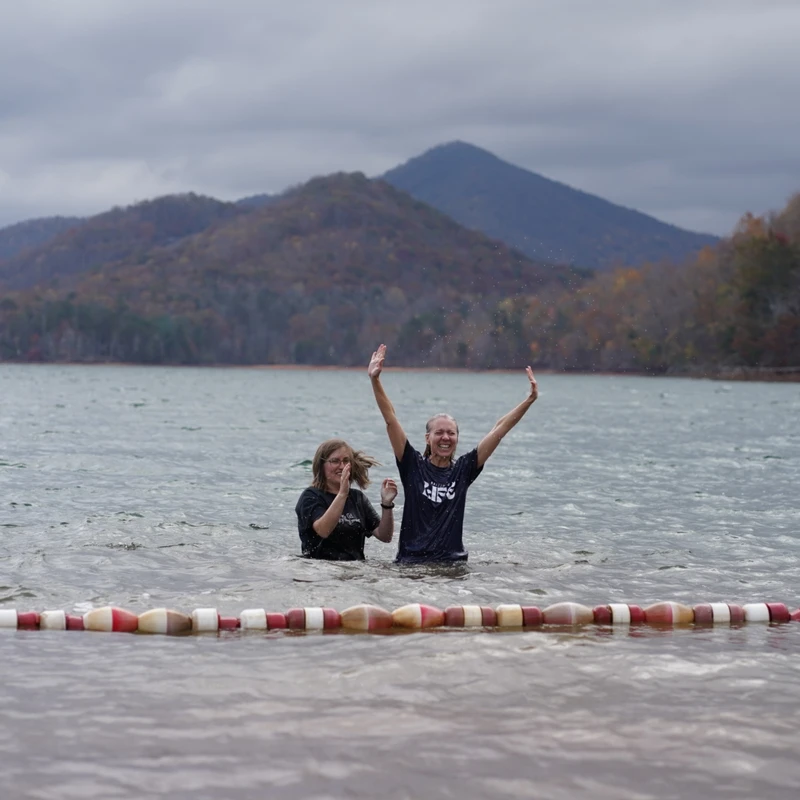 Two women stand waist-deep in a lake after a baptism. One raises her hands in celebration while the other claps, with mountains in the background.