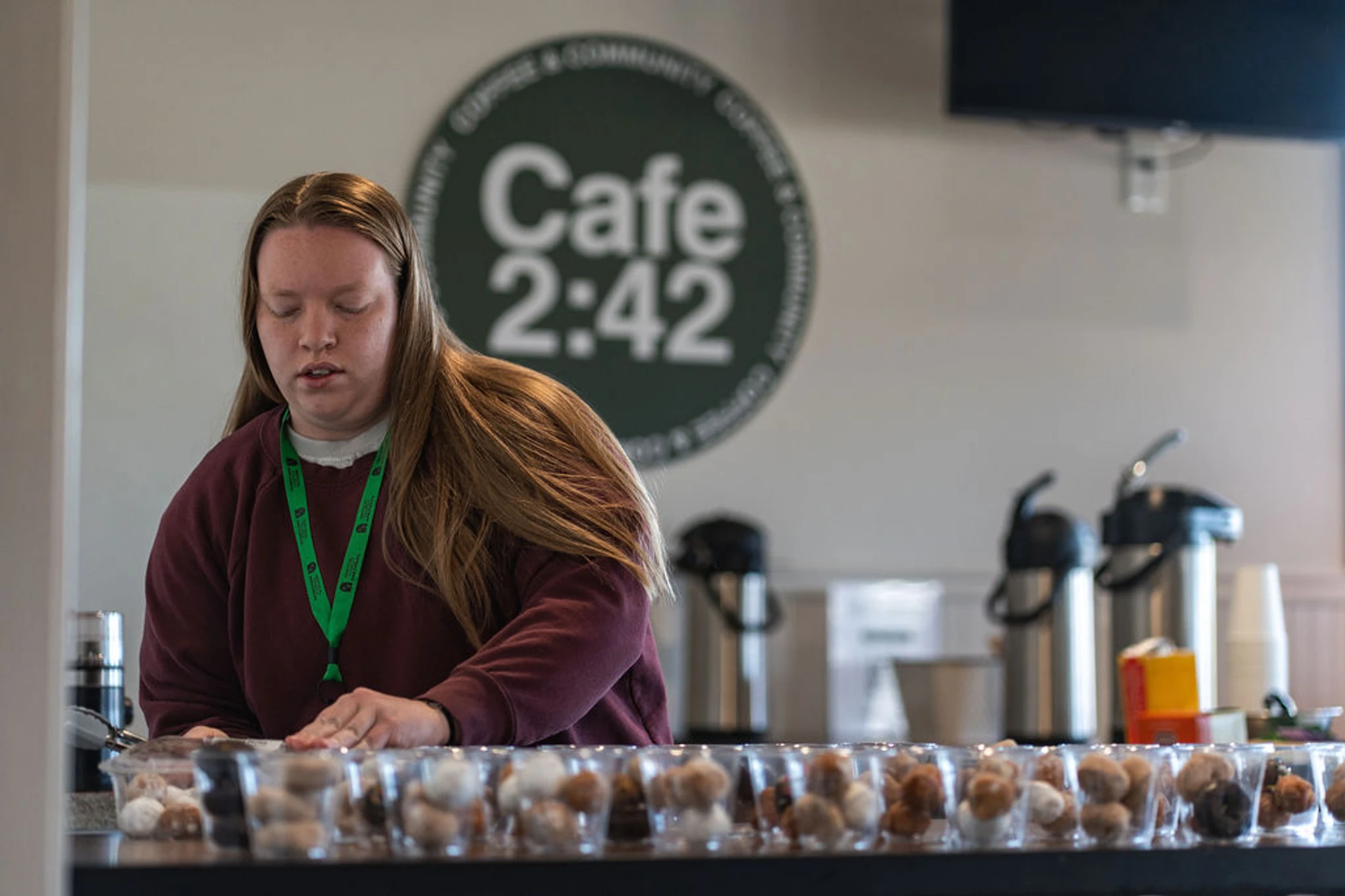 A young woman in a maroon sweatshirt stands behind a counter at Cafe 2:42, arranging clear plastic cups filled with assorted donut holes.