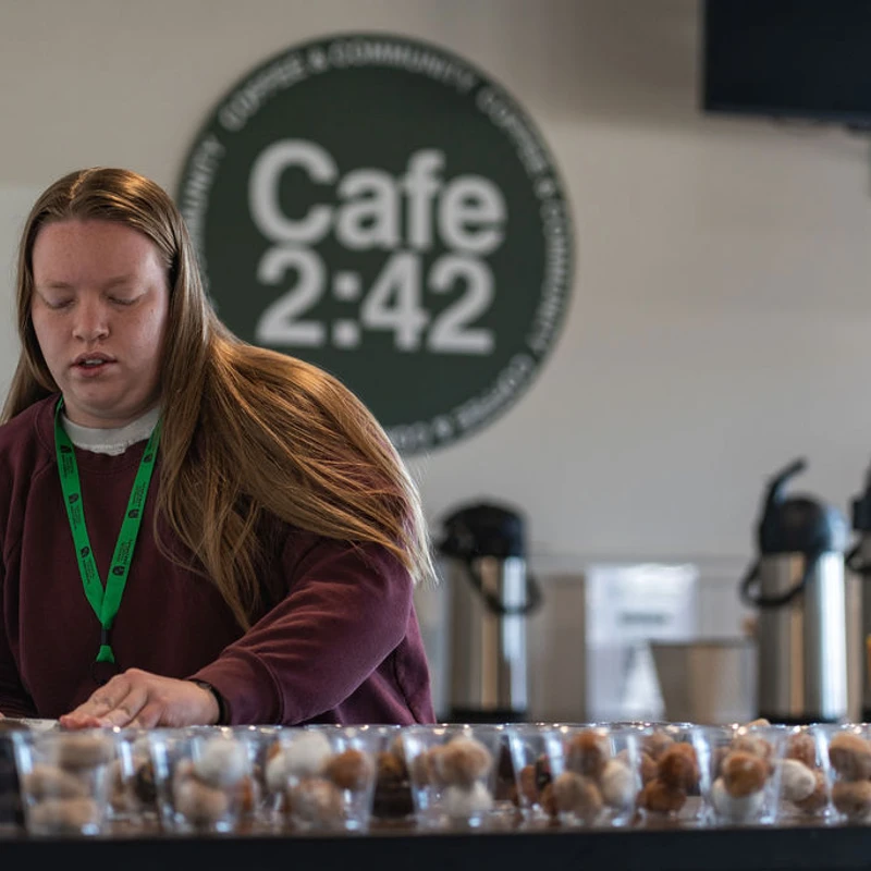 A young woman in a maroon sweatshirt stands behind a counter at Cafe 2:42, arranging clear plastic cups filled with assorted donut holes.