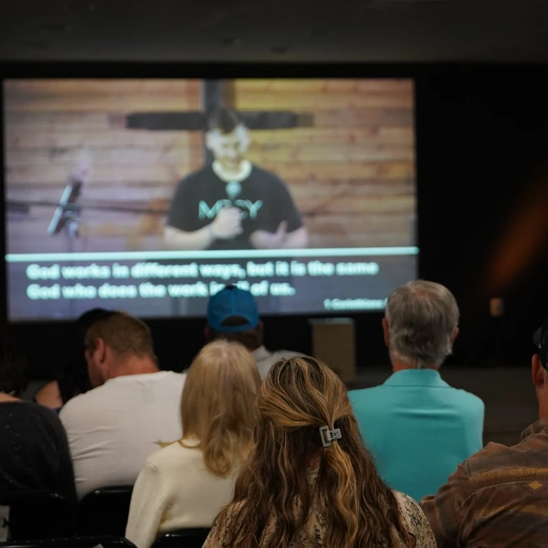 The backs of an audience sitting in chairs watching a man give a sermon on a large screen. The screen has subtitles and a large cross is visible.