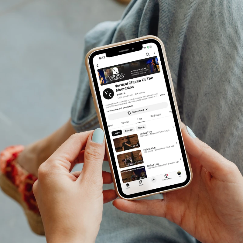 A person with light blue nails holds a smartphone, viewing the YouTube channel for Vertical Church Of The Mountains and its live stream videos.