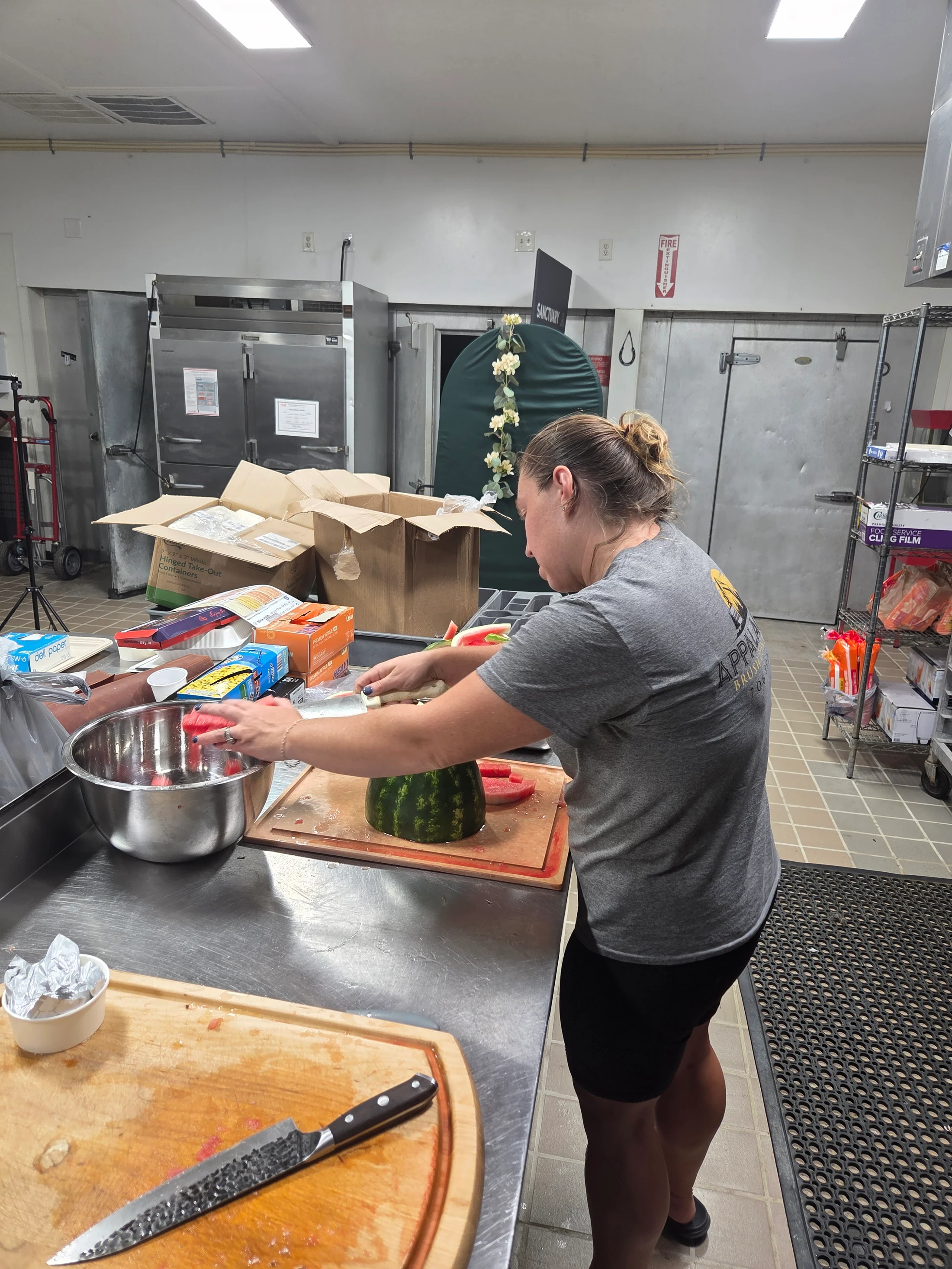 A woman with her hair in a bun stands in a commercial kitchen, slicing a large watermelon on a cutting board and placing the pieces into a metal bowl.