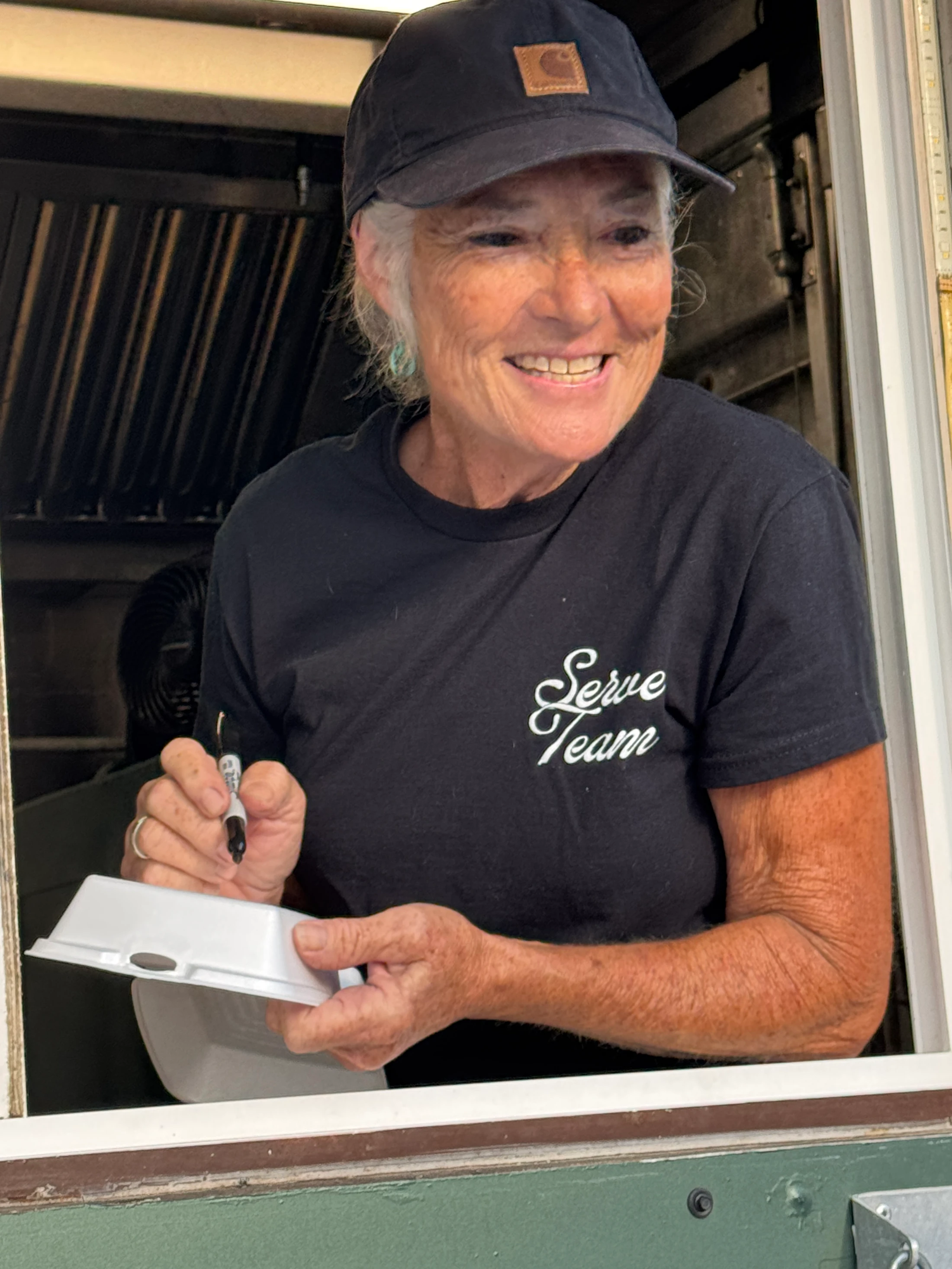 A smiling woman with gray hair wears a black 'Serve Team' t-shirt and a Carhartt cap. She leans out of a service window, writing on a styrofoam to-go box.