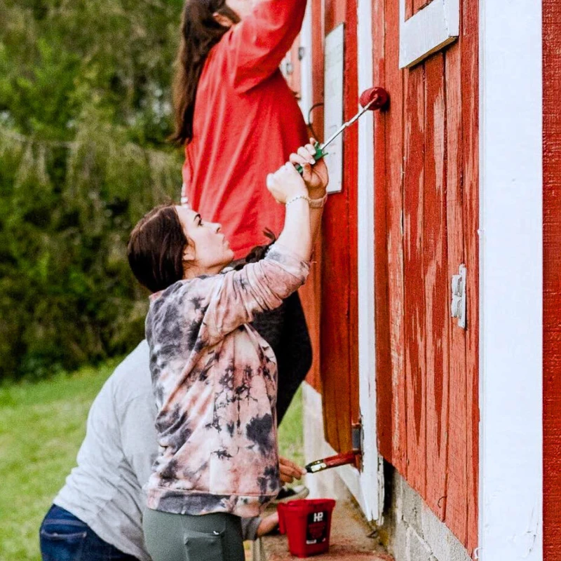 Three people work together painting the side of a red wooden barn. A woman in a tie-dye shirt uses a roller while a man in a red shirt paints above her.