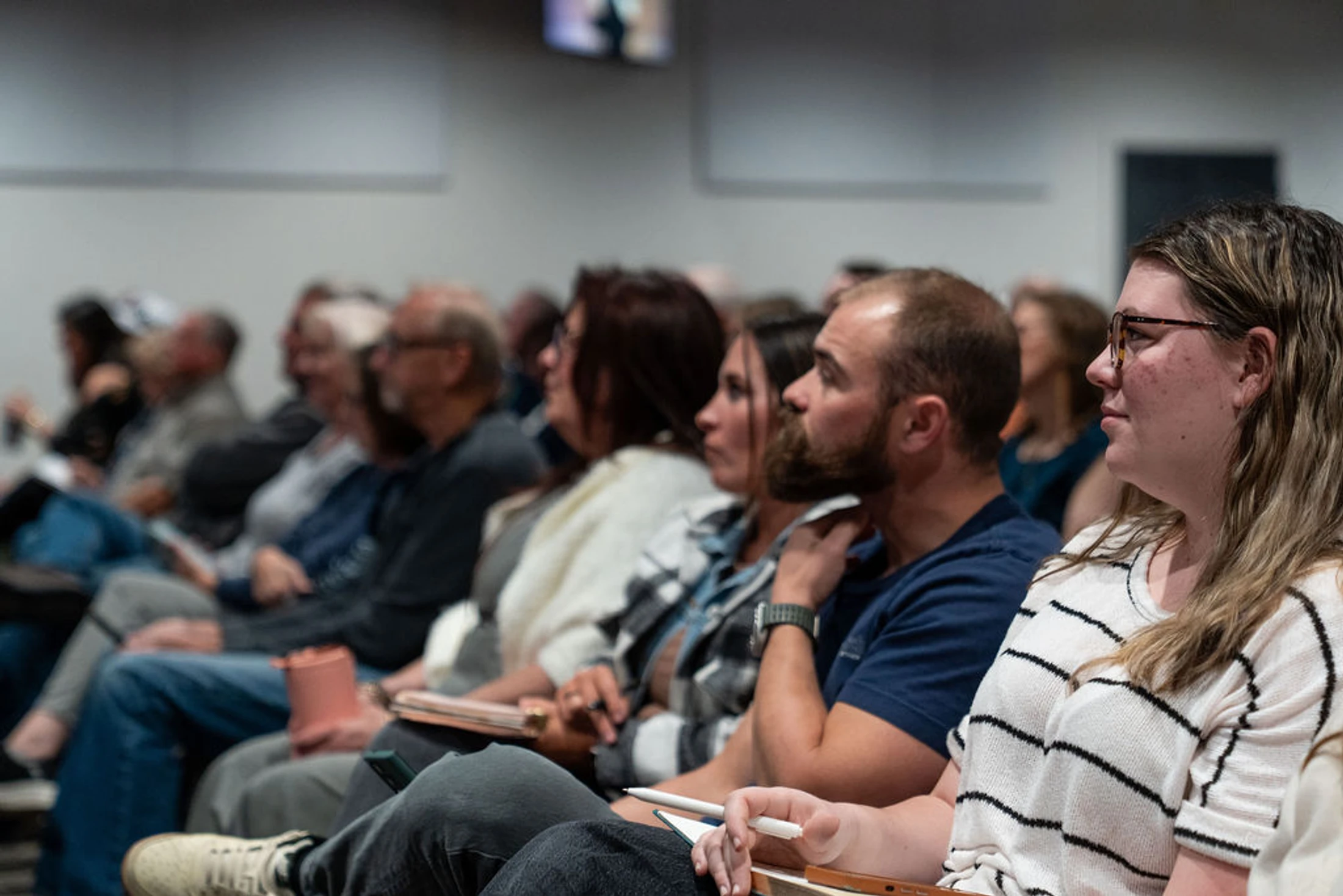A side-profile view of people sitting in rows as an audience. The focus is on a woman with glasses and a man with a beard in the foreground.