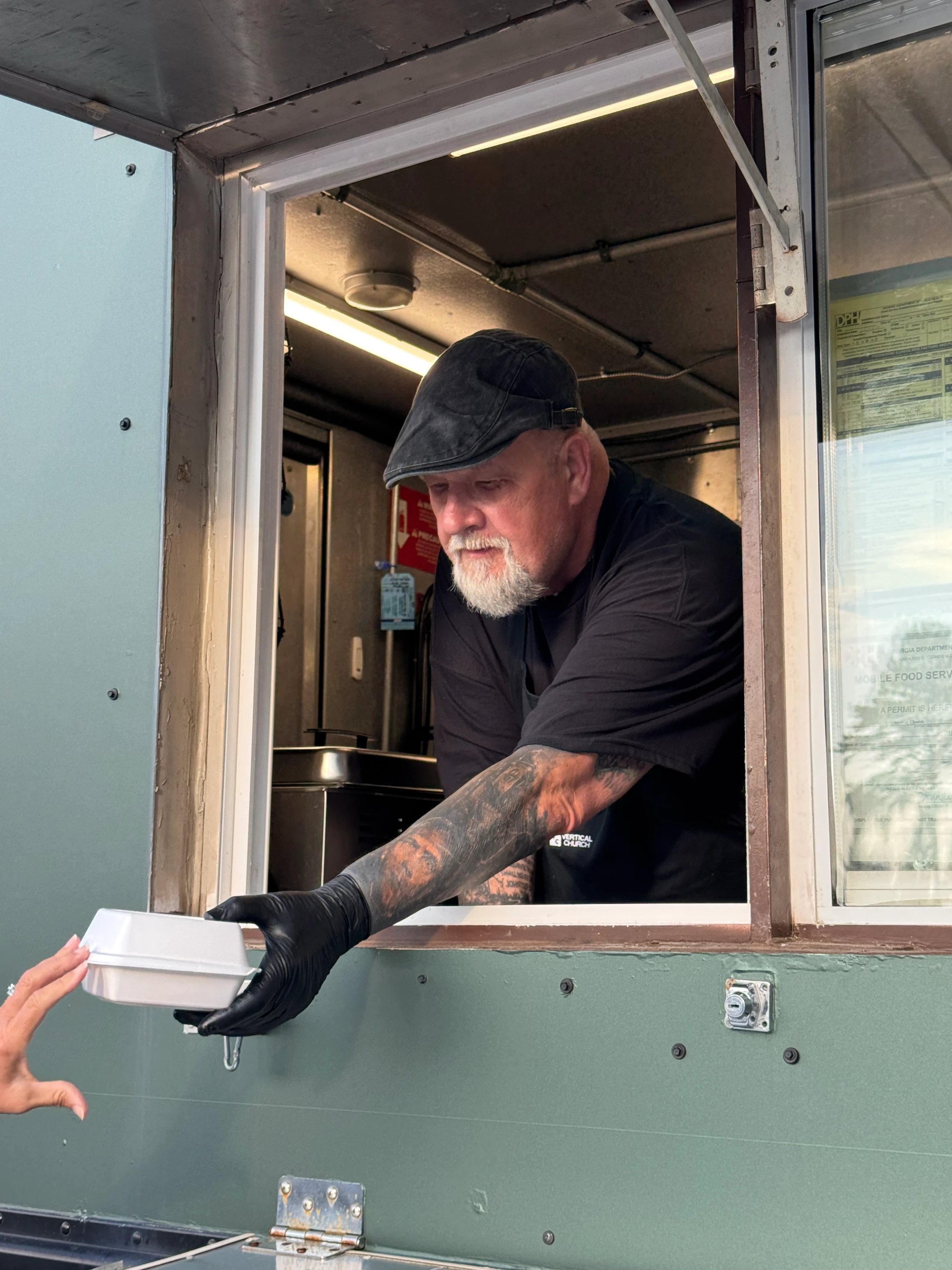 A man with a goatee and tattoos wearing a flat cap and gloves hands a takeout container to a customer from a food truck service window.