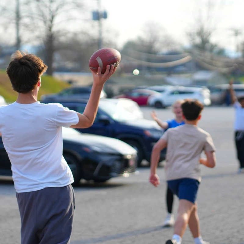 A group of teenagers plays a casual game of football in a parking lot. The focus is on a boy in a white shirt from behind, about to throw the ball.