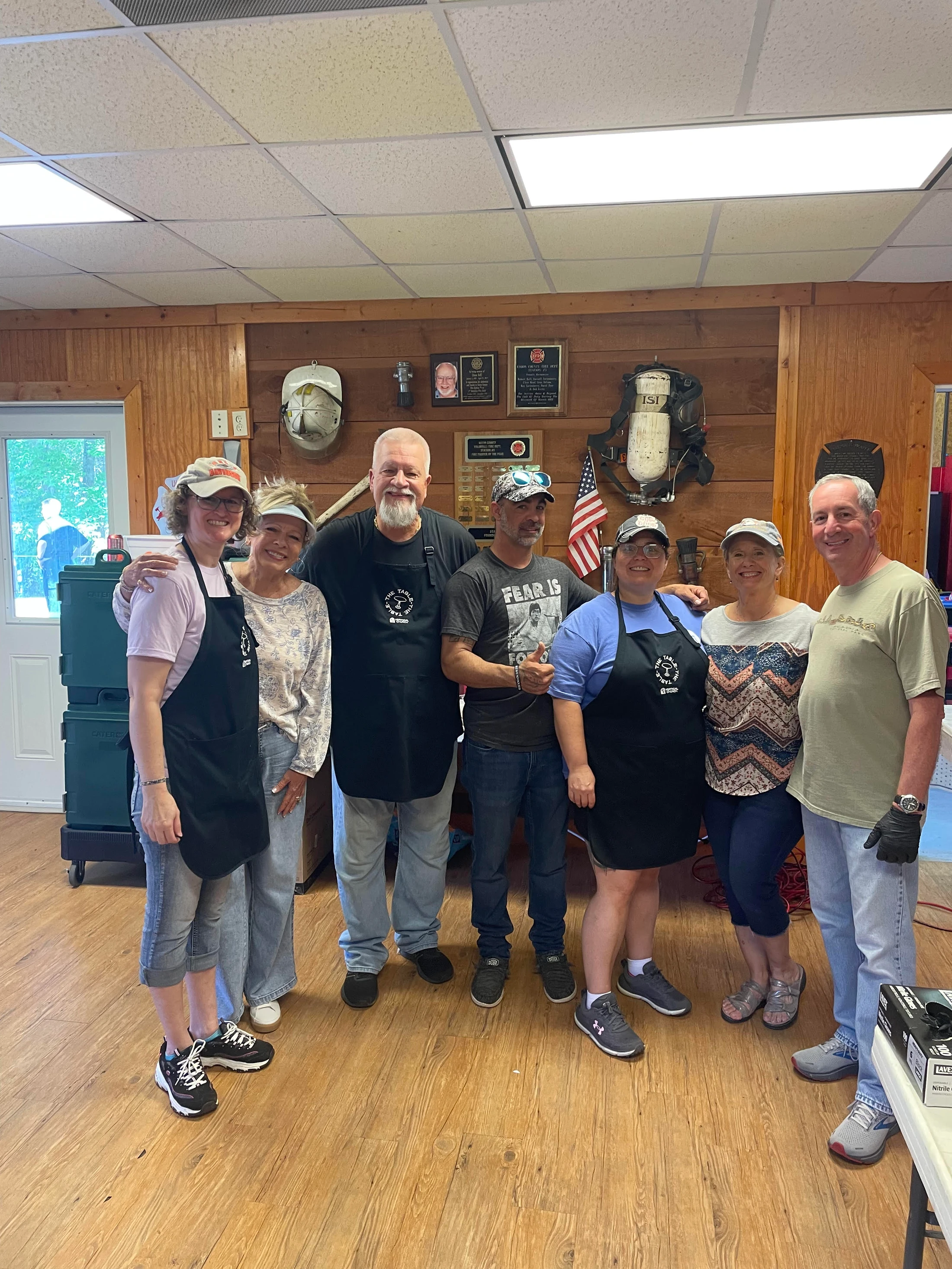 A group of seven men and women in aprons and casual wear smile for the camera while standing in a room with wood-paneled walls decorated with plaques.