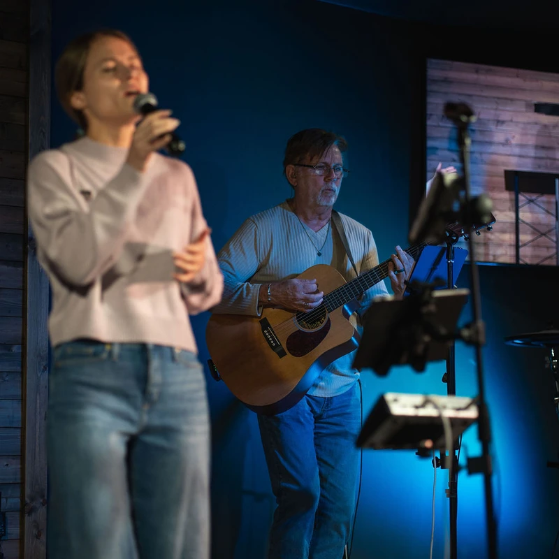 A woman sings into a microphone in the foreground while a man in the background plays an acoustic guitar on a dimly lit stage with blue lighting.