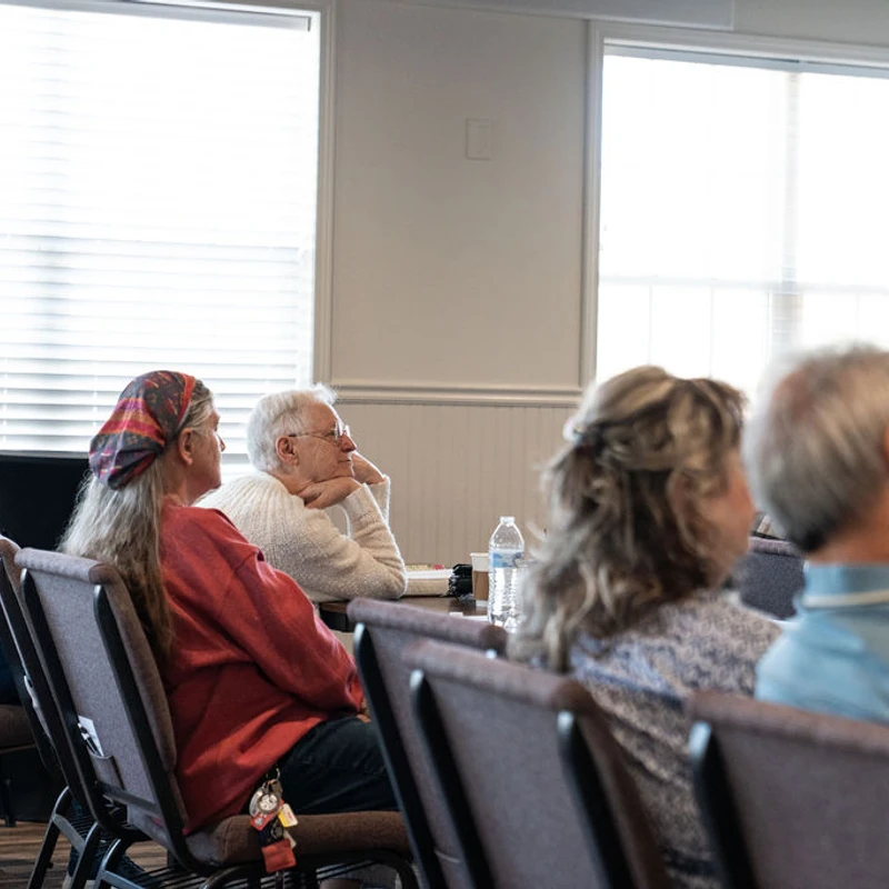 A side view of a church congregation sitting in rows of chairs, listening attentively during a service. A 'Vertical Church' banner is visible in the background.