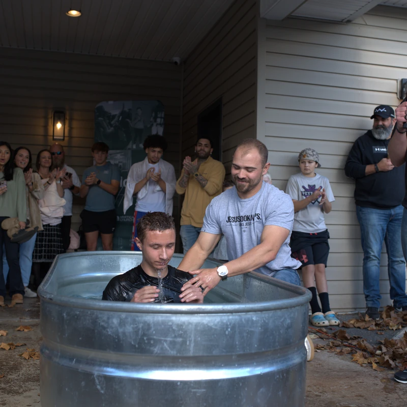A young man is baptized in a large metal tub on a patio as a crowd of people watch, clap, and smile. A man with a microphone officiates the ceremony.