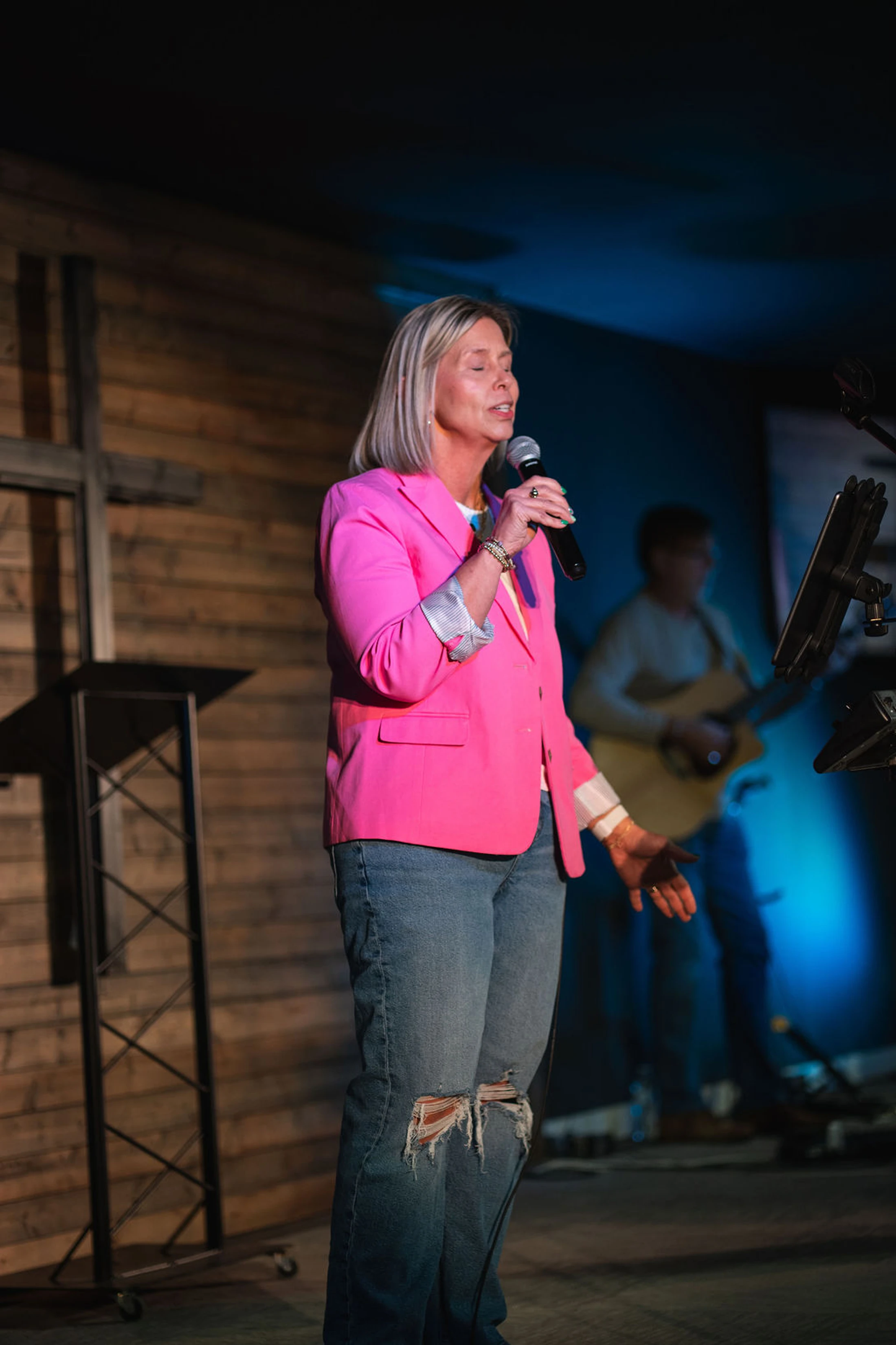 A woman with blonde hair in a bright pink blazer and ripped jeans sings into a microphone on a dimly lit stage with a wooden wall and cross behind her.