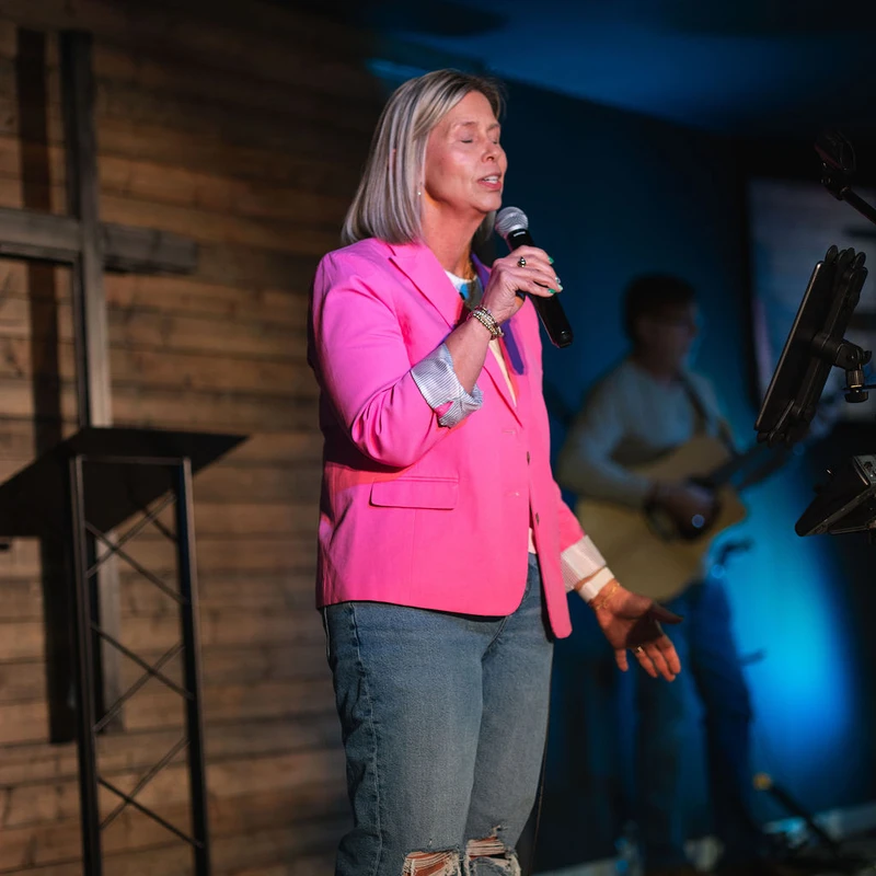 A woman with blonde hair in a bright pink blazer and ripped jeans sings into a microphone on a dimly lit stage with a wooden wall and cross behind her.
