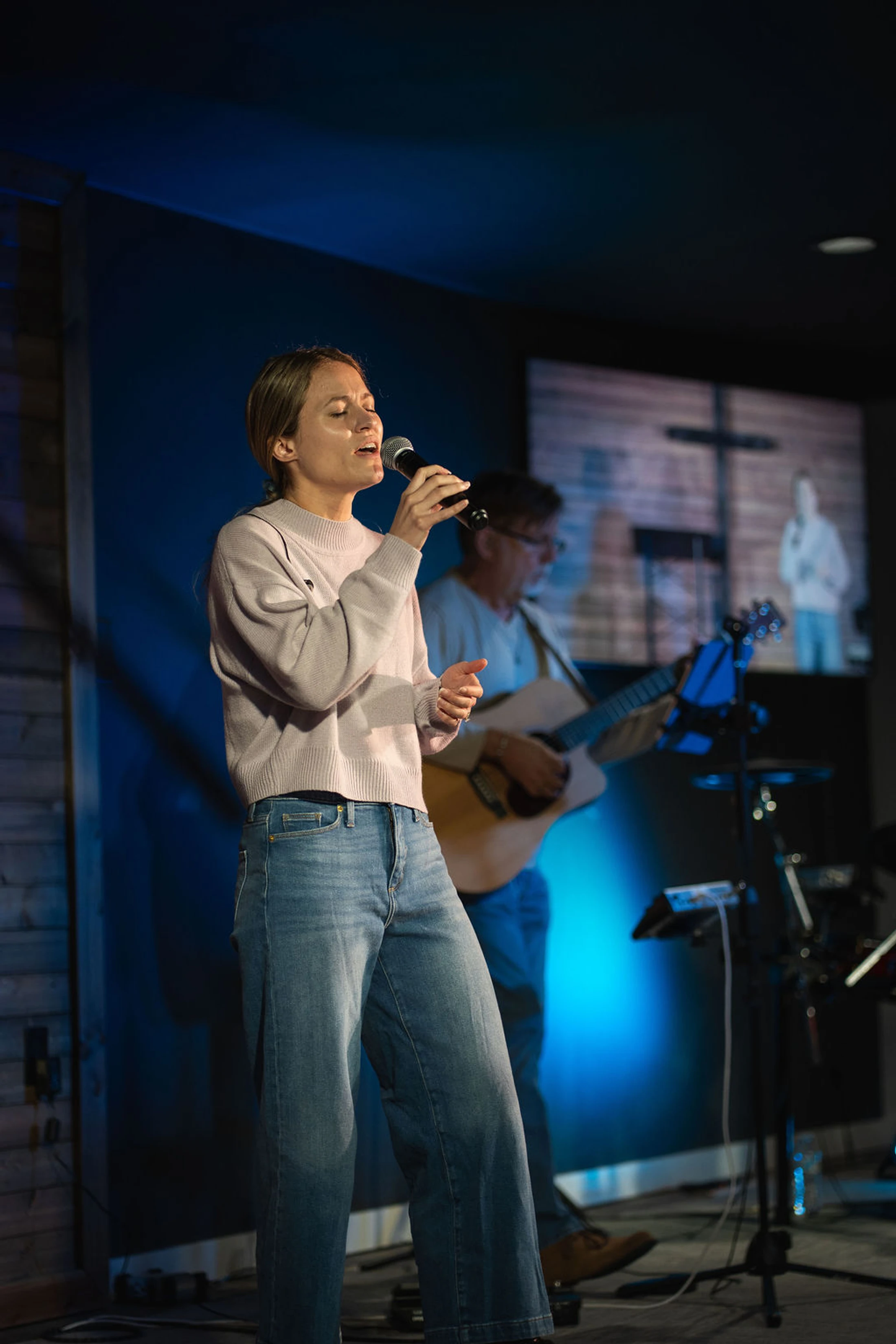 A woman in a lavender sweater and jeans sings into a microphone on a dimly lit stage with blue lights. A man playing guitar is visible behind her.