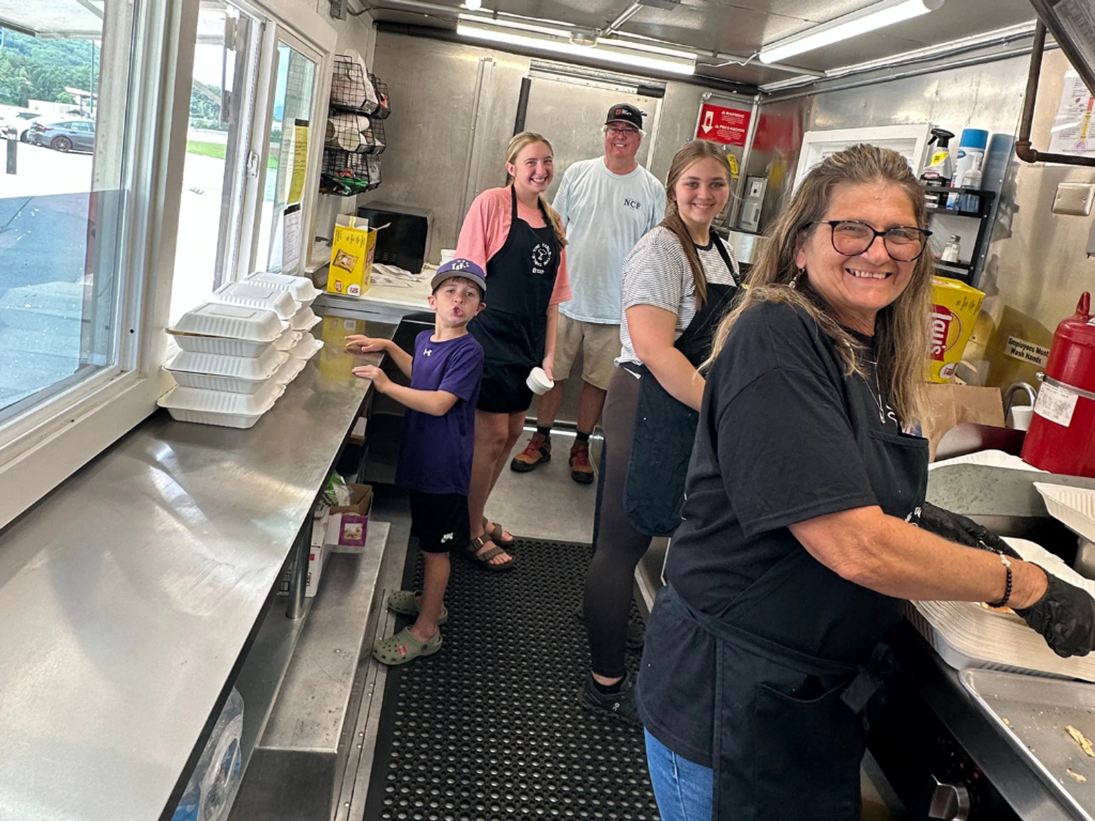 A smiling family of five, including a young boy, poses for a photo while working inside a busy food truck with stainless steel counters.