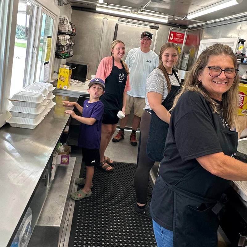 A smiling family of five, including a young boy, poses for a photo while working inside a busy food truck with stainless steel counters.