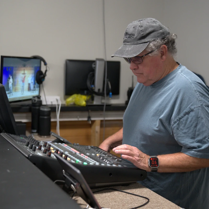 A man in a hat and glasses stands at a large soundboard, adjusting the controls. A monitor in the background shows a live church service.