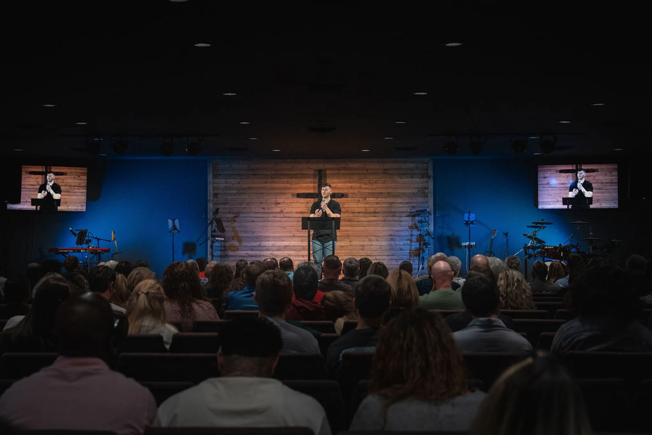A pastor stands on a warmly lit stage with a wooden wall and cross, speaking to a large congregation seated in a dimly lit auditorium.