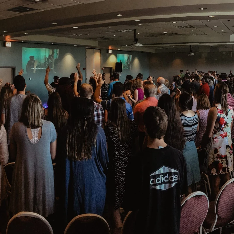 A wide-angle photo from the back of a crowded room showing people standing and raising their hands in worship, facing screens at the front.