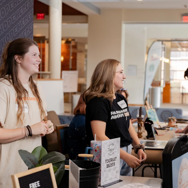 Two young women smile as they stand behind a welcome table at a college ministry event, with a sign that reads 'FREE ICED COFFEE' in the foreground.