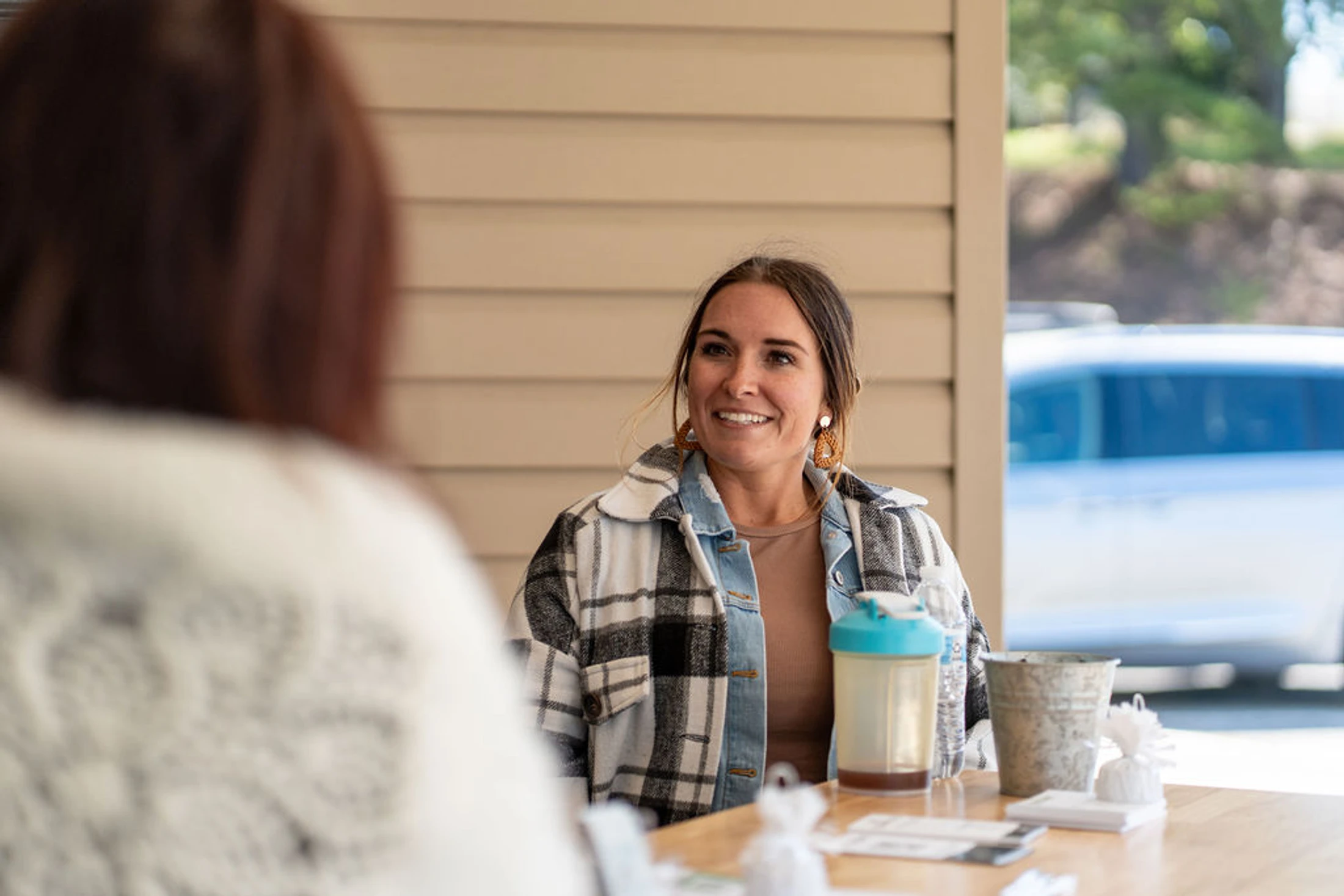 A woman with brown hair smiles while sitting at a wooden table outdoors, engaged in a conversation with another person whose back is to the camera.