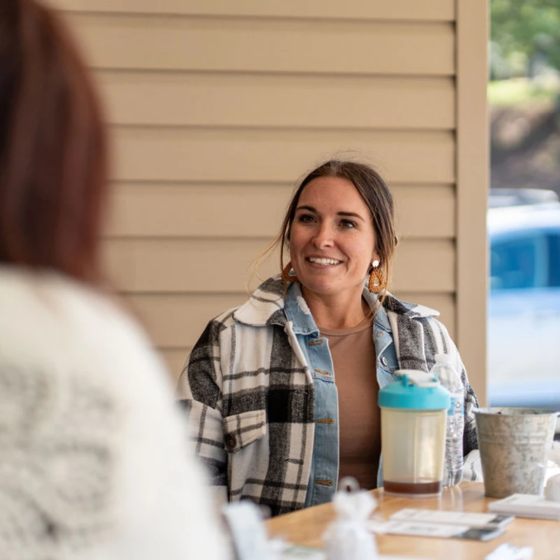 A woman with brown hair smiles while sitting at a wooden table outdoors, engaged in a conversation with another person whose back is to the camera.